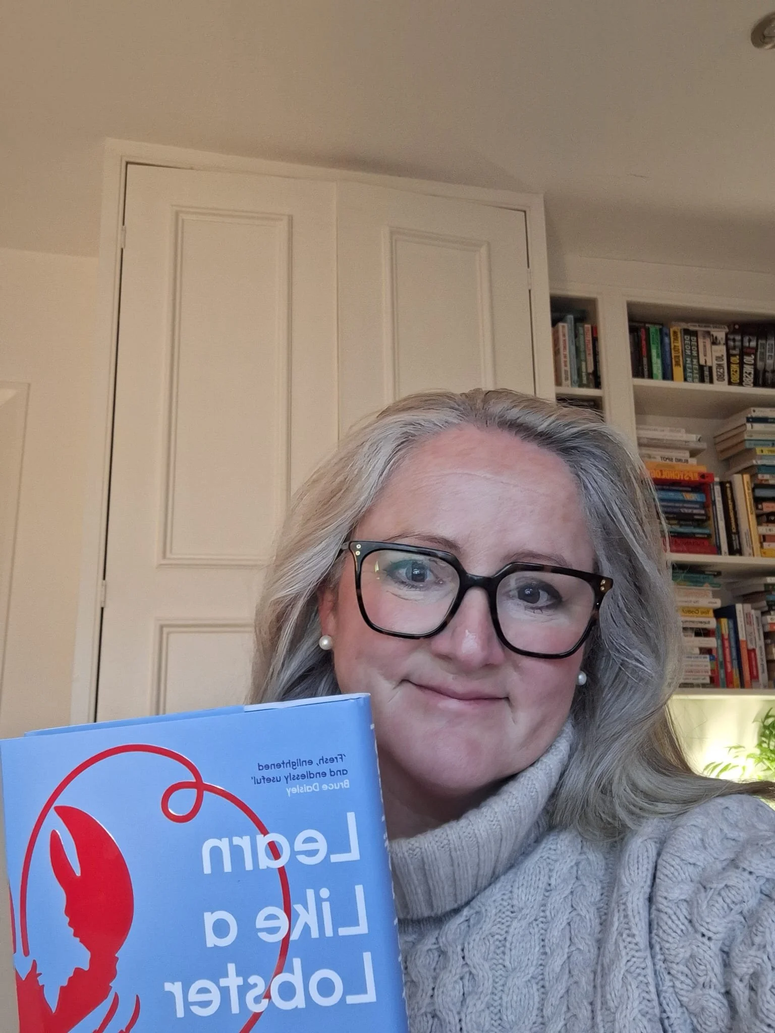 A woman with gray hair, glasses, and pearl earrings holding a blue book in front of bookshelves and a closet door.