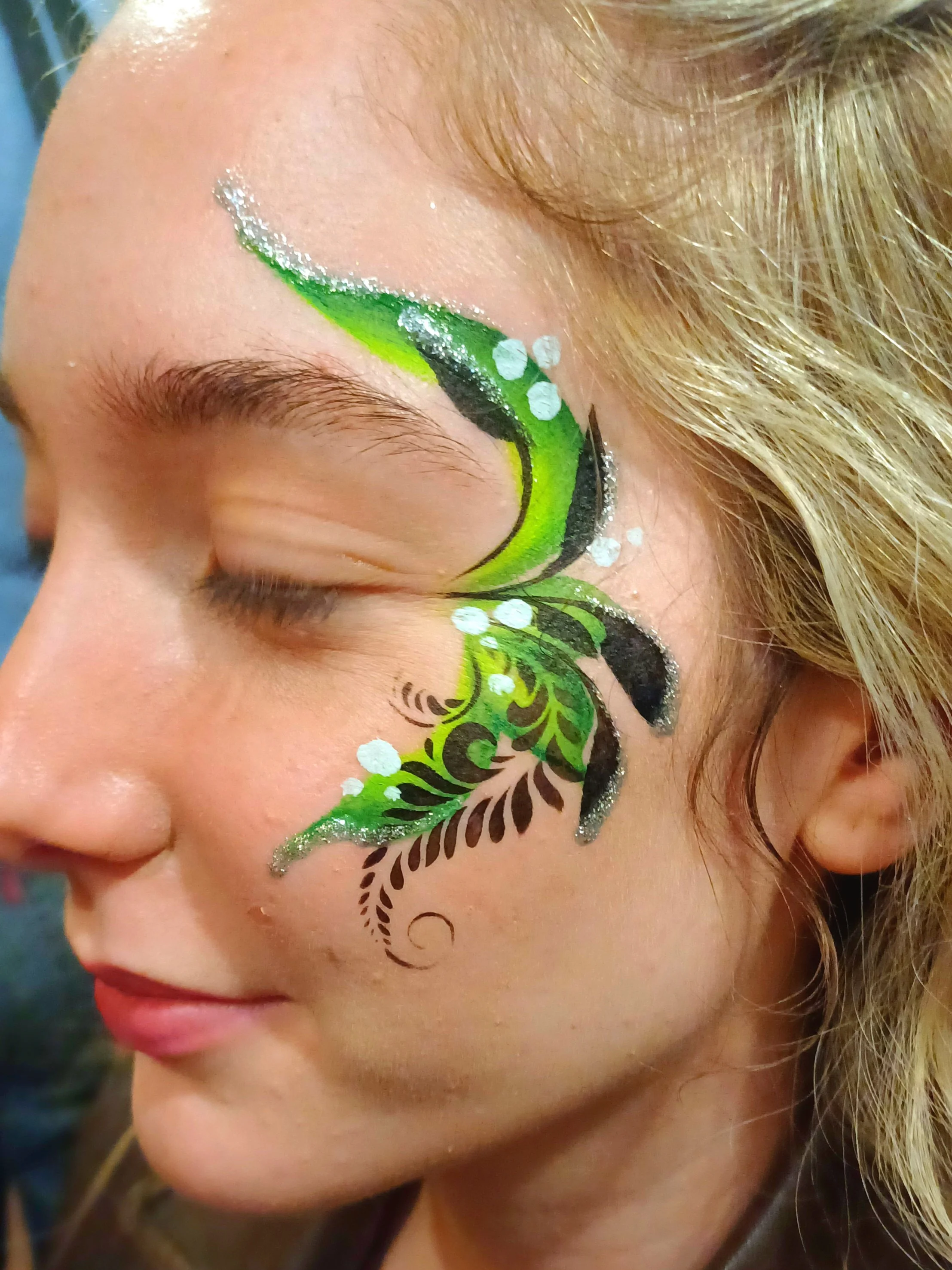 Close-up of a woman's face with elaborate face paint in green, black, and white, accented with glitter, covering the eye and cheek area.