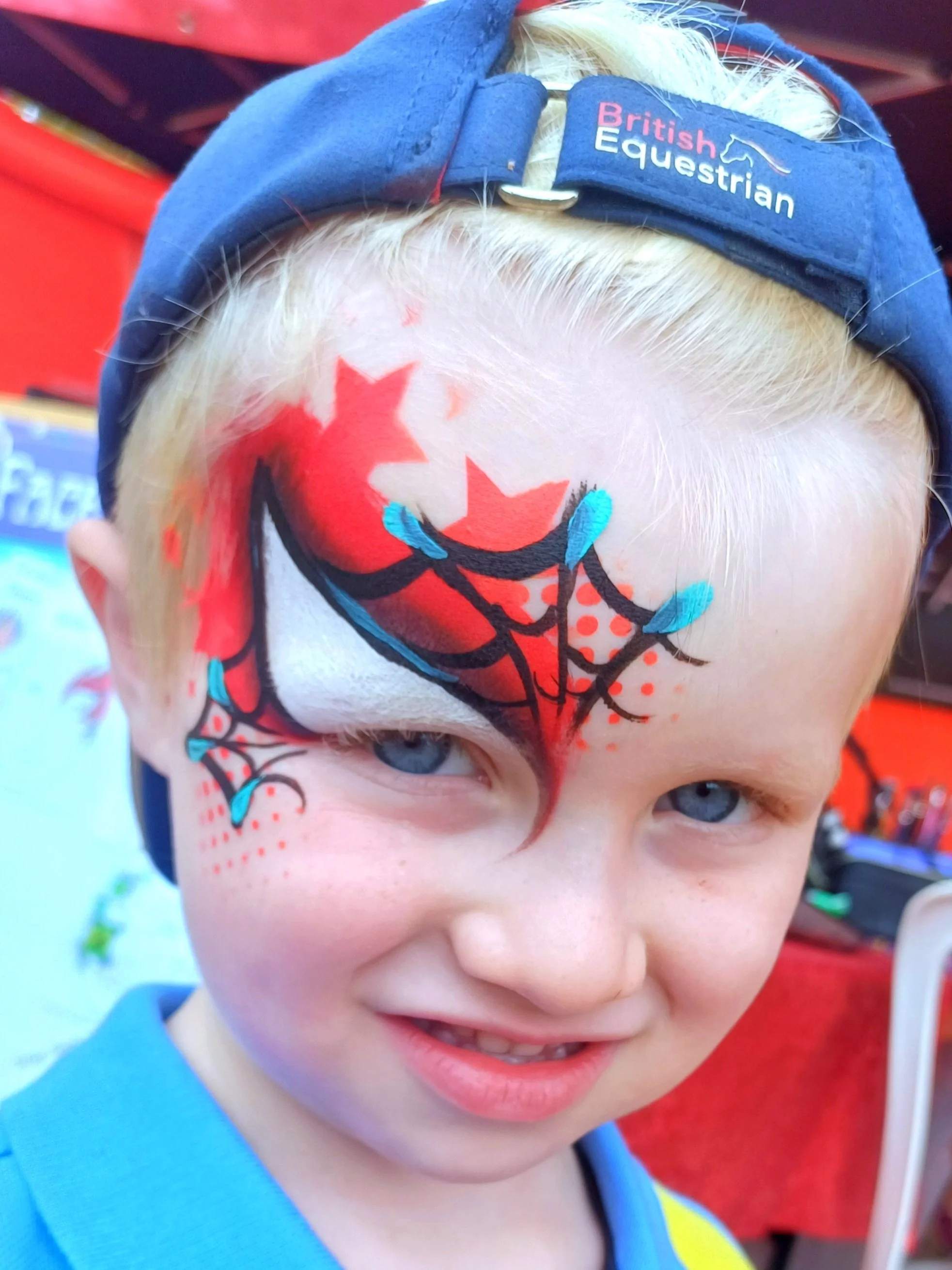 Close-up of a young boy with blonde hair wearing a blue cap with a 'British Equestrian' logo, and face paint featuring red stars, black web, and blue accents on his forehead, under a red tent.