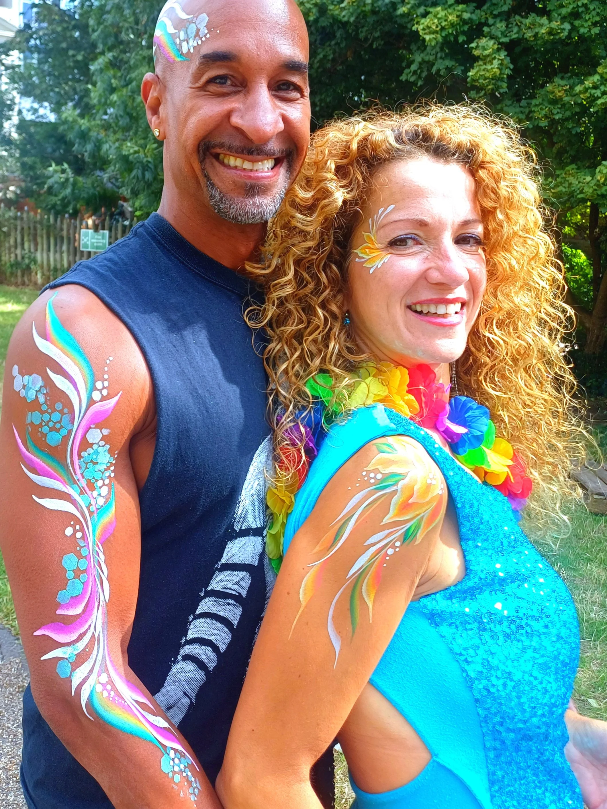 Two people with painted bodies and faces, one man and one woman, smiling outdoors. The man has a rainbow and white design on his bald head and arms, and the woman has a flower face paint and curly hair, wearing a bright blue dress and a rainbow lei. Background includes trees and a wooden fence.