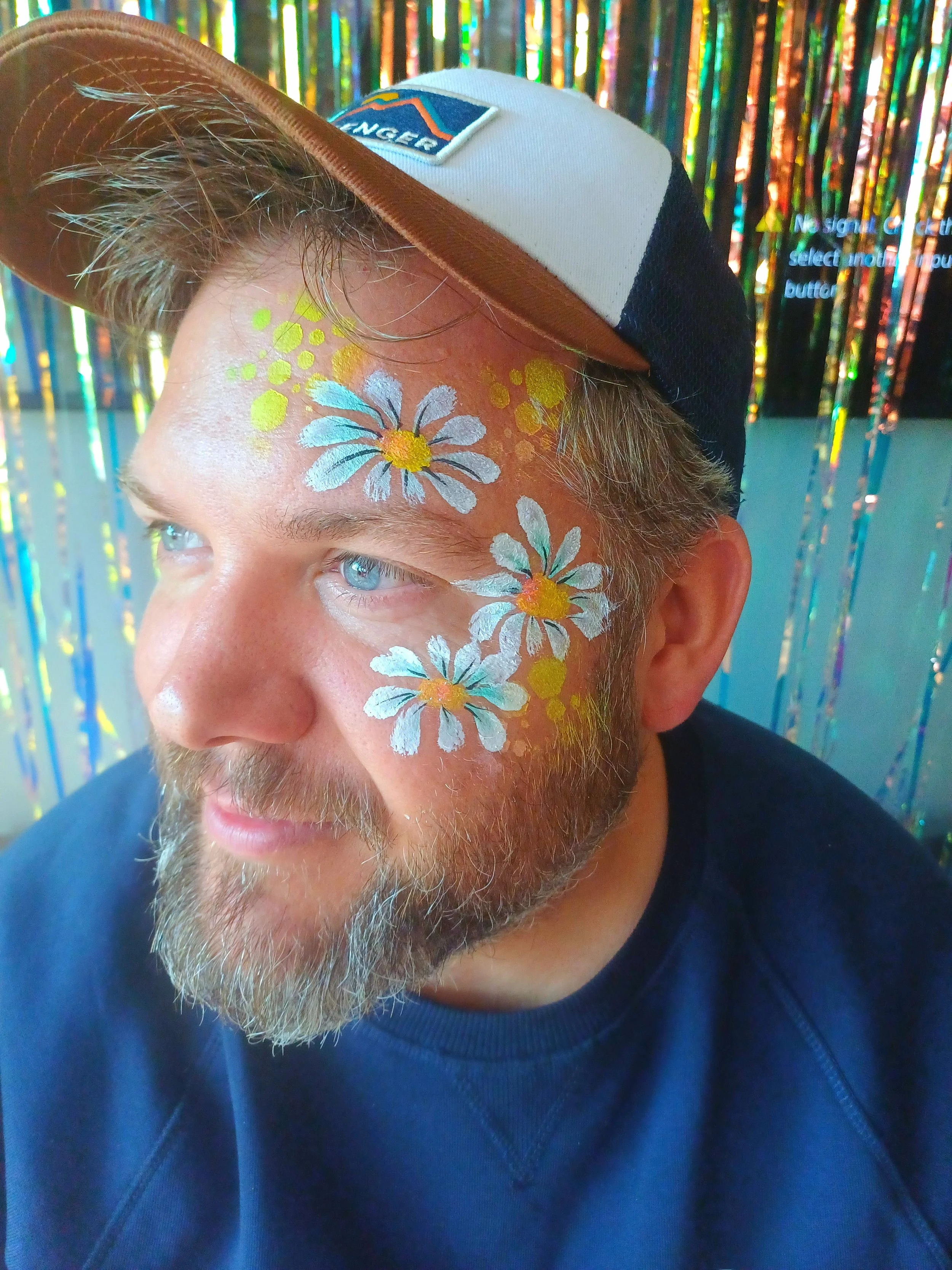 Close-up of a man's face with painted flowers and yellow circles, wearing a two-tone cap with a patch, and a blue shirt, against a shiny, colorful background.