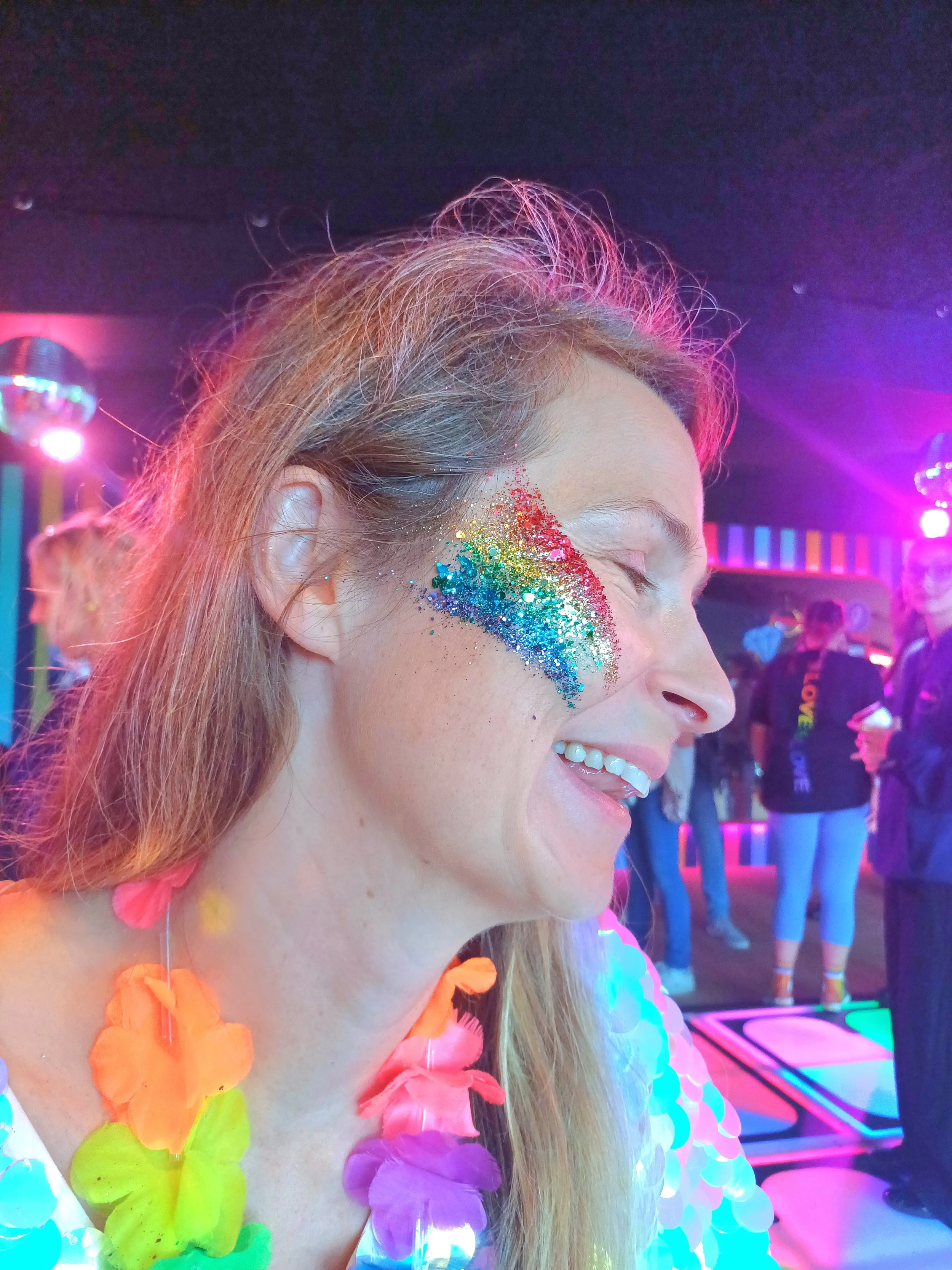 A woman with long hair and colorful glitter makeup on her cheek, wearing a neon-colored lei and a dress with pastel ruffles, smiling with eyes closed at a neon-lit party or event.