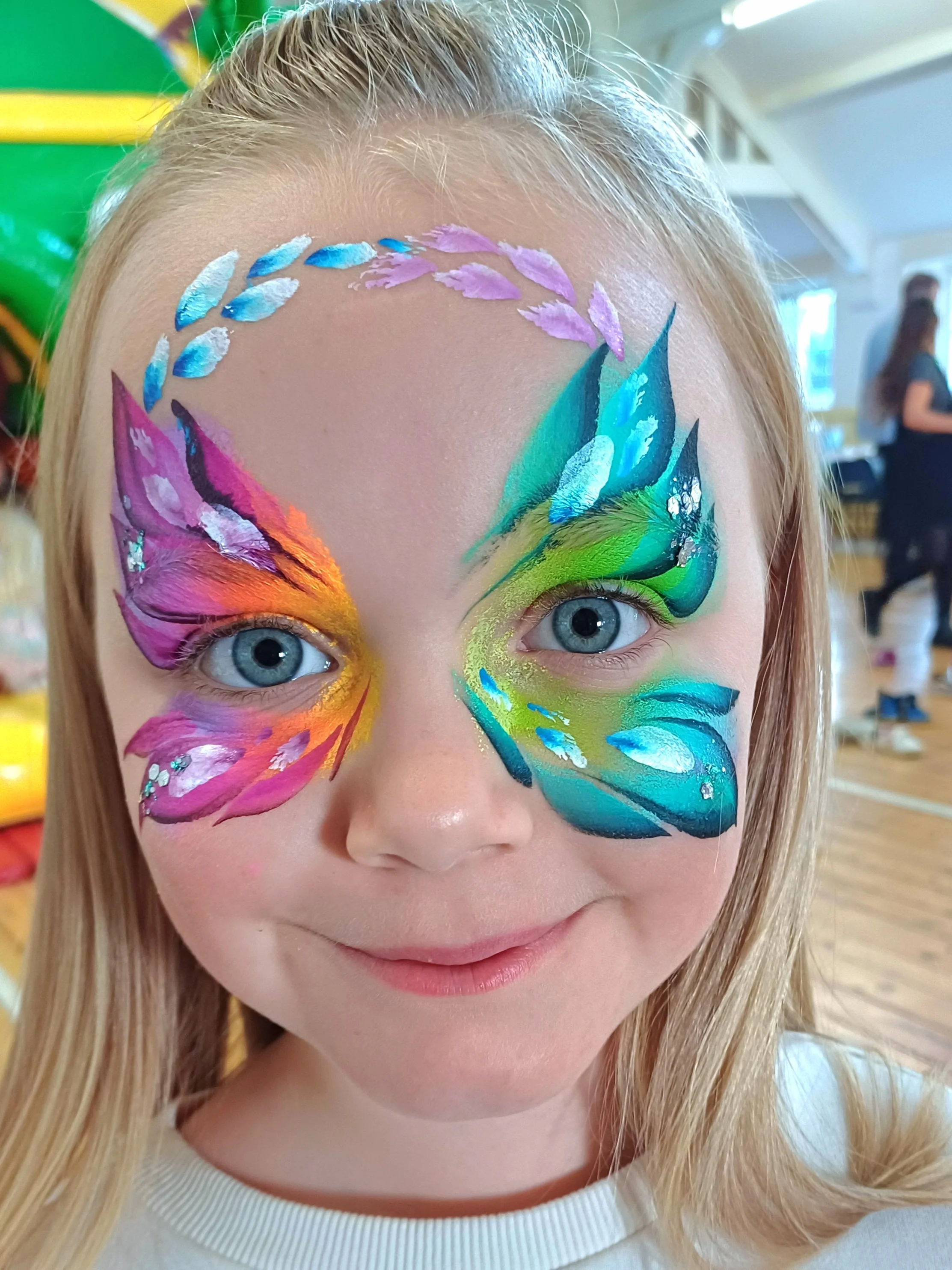 Close-up of a young girl with intricate, colorful butterfly face paint covering her eyes, cheeks, and forehead. The face paint includes shades of blue, purple, green, yellow, pink, and white, creating a vibrant butterfly design.