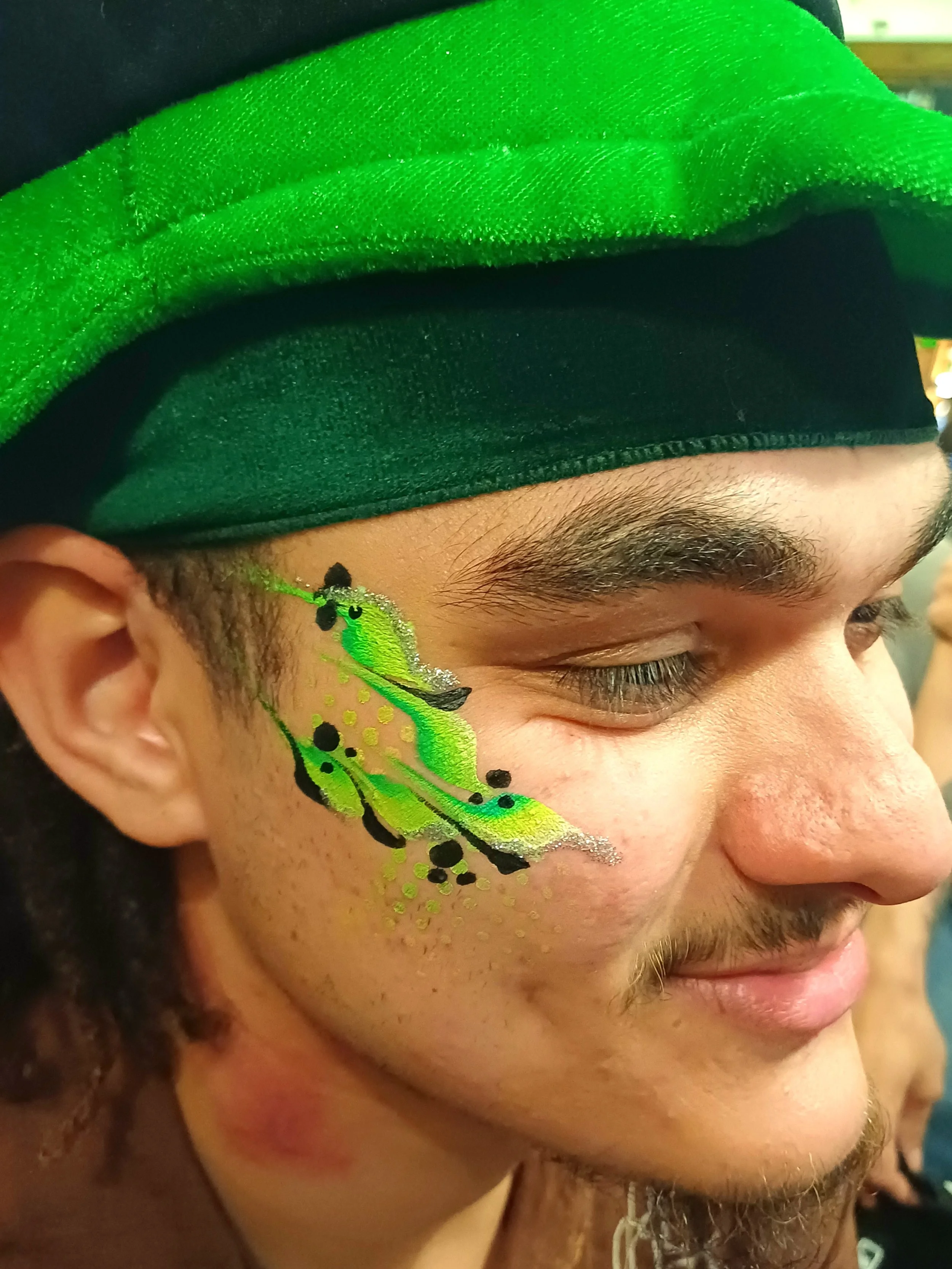 Close-up of a person with detailed, colorful face paint in green, black, and yellow, and wearing a black and green hat.