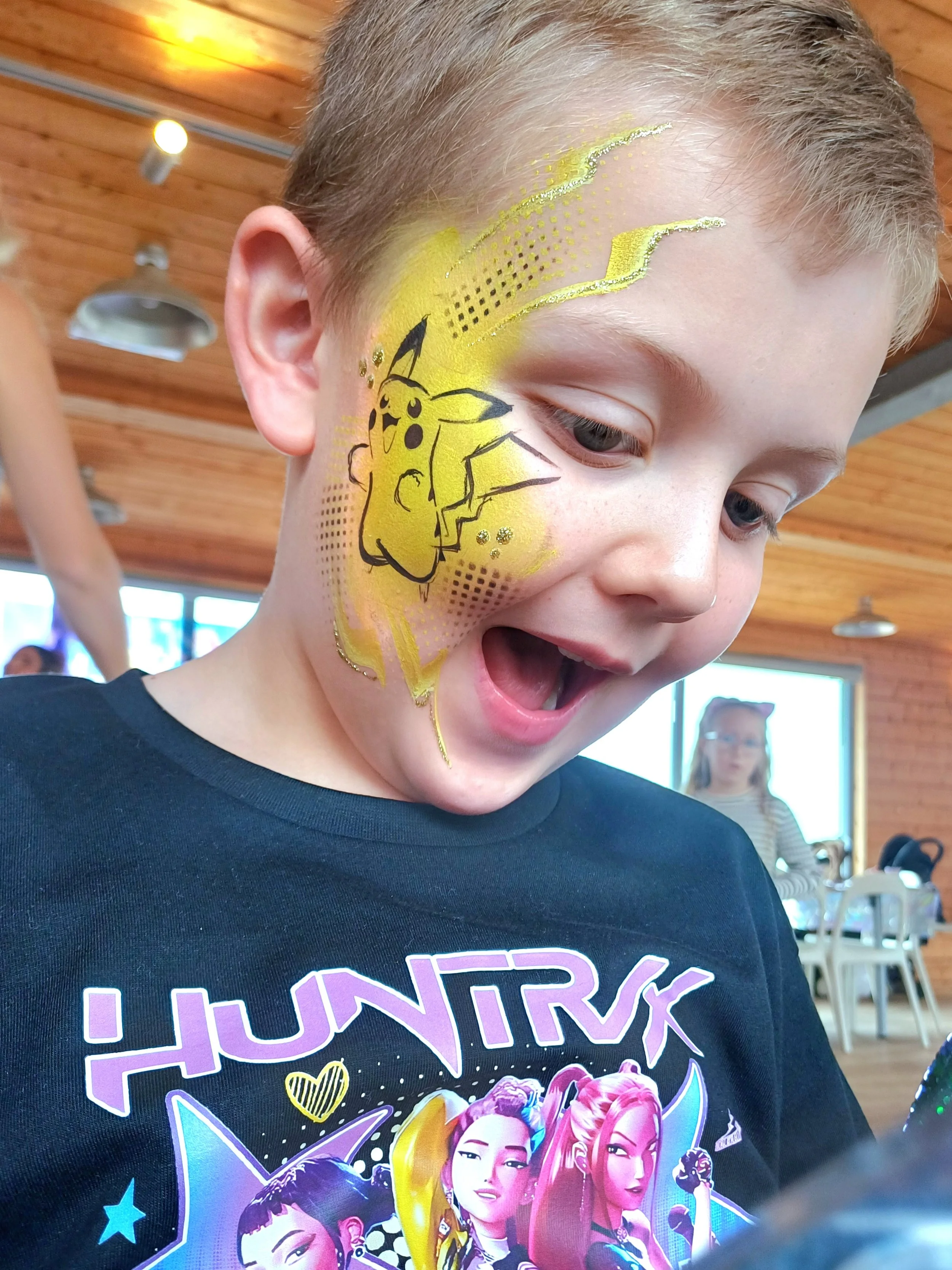 Close-up of a boy with face paint of Pikachu from Pokémon, wearing a black t-shirt with multicolored graphics, in an indoor setting with wooden ceiling and a woman in the background.