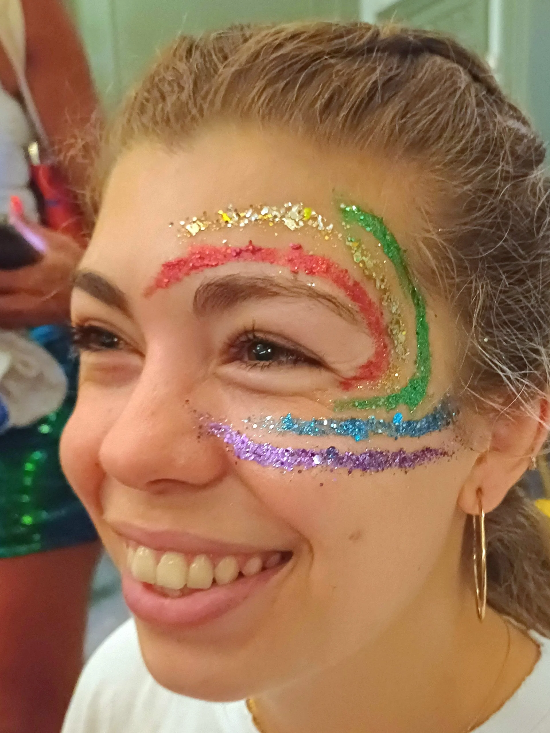 Close-up of a smiling woman with colorful glitter face paint and makeup around her right eye, featuring red, green, yellow, blue, and purple glitter, with her hair styled back and a gold hoop earring visible.