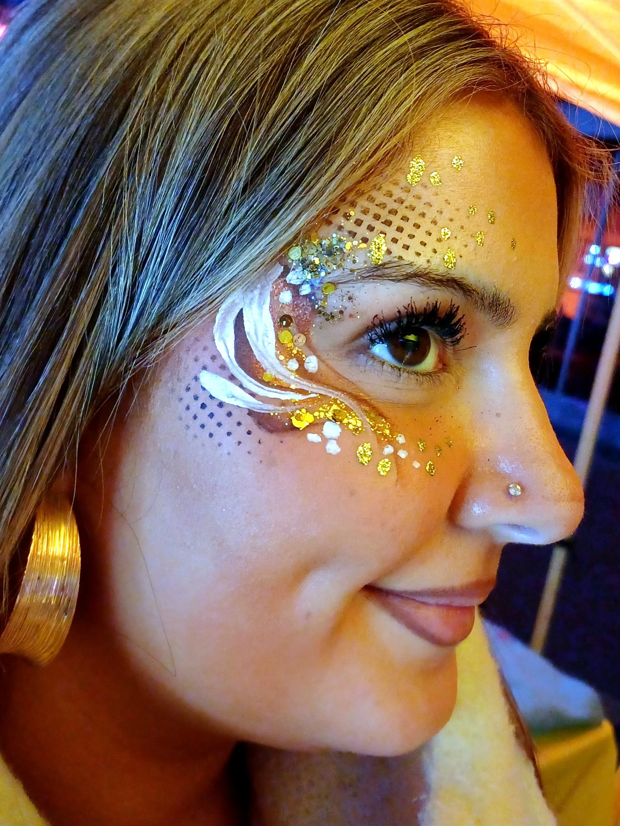 Close-up of a woman's face with gold glitter and white face paint surrounding her eye.