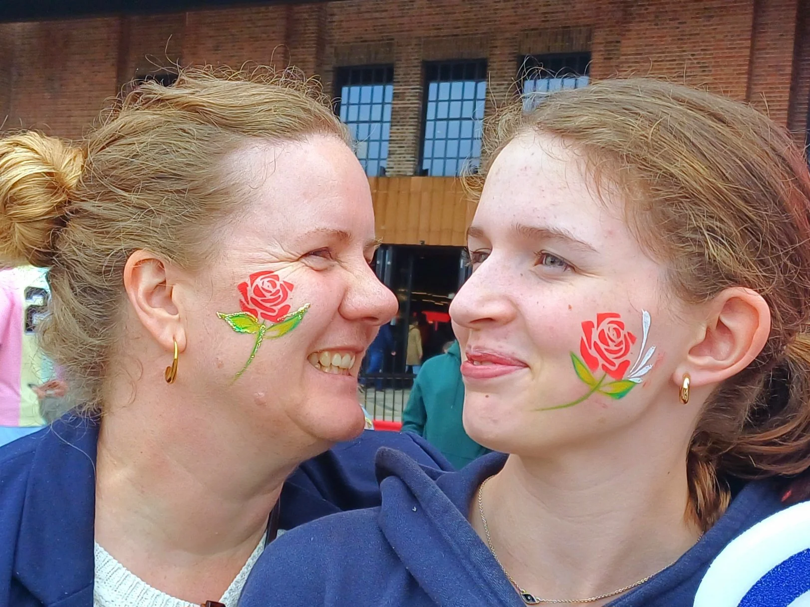 Two women smiling at each other, with painted red roses and green leaves on their cheeks, standing outdoors in front of a brick building with windows.