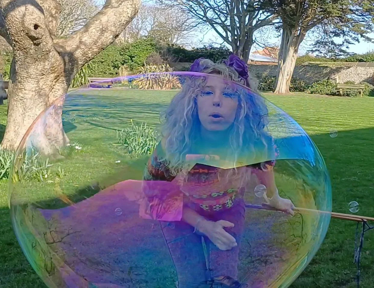 A woman with curly blonde hair and purple accessories blowing a large soap bubble outdoors in a grassy yard with trees and a stone wall.