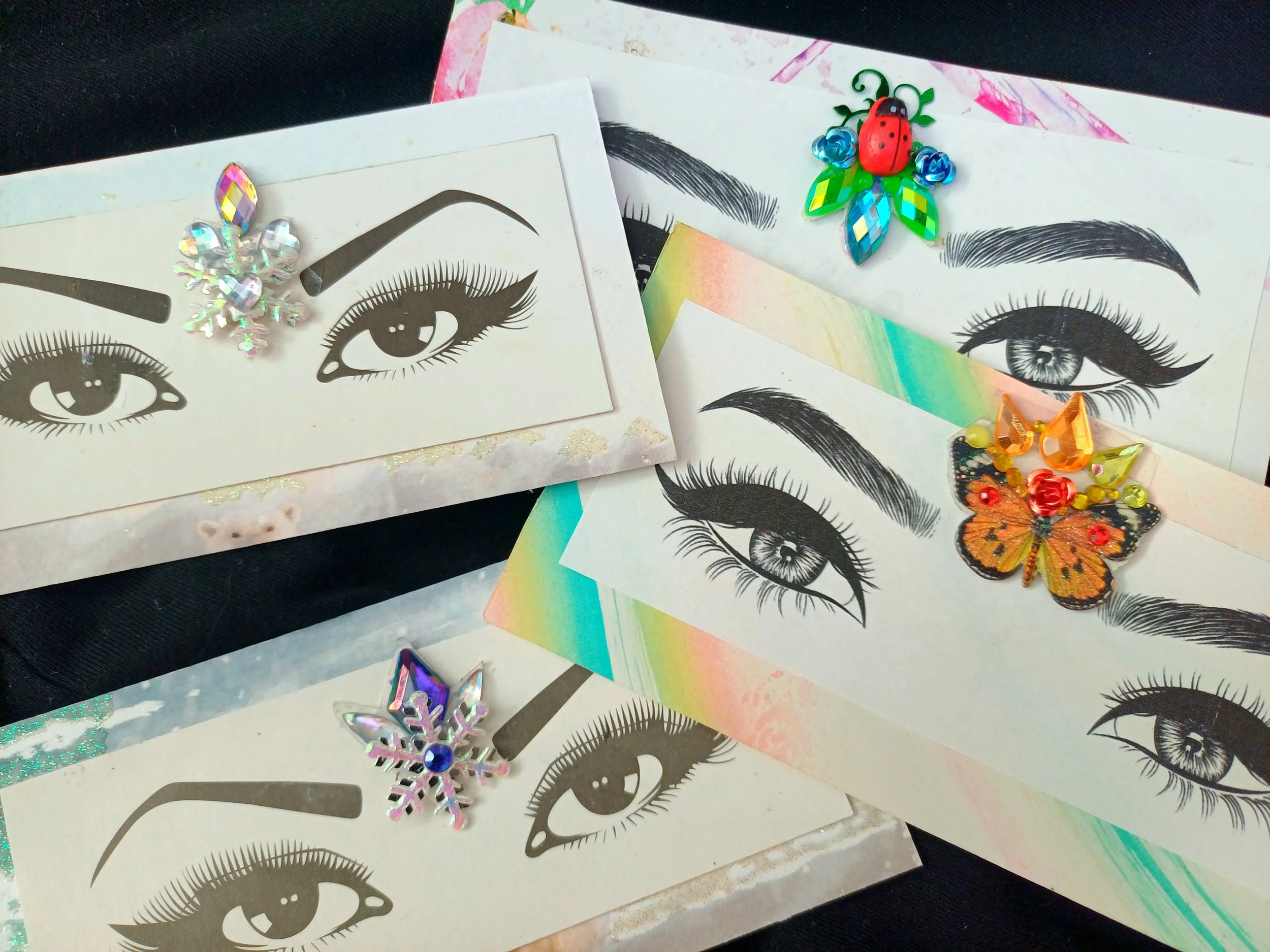 Assorted decorative stickers and embellishments featuring eyes with long lashes, a ladybug, a butterfly, snowflake, and colorful embellishments on sheets of paper.
