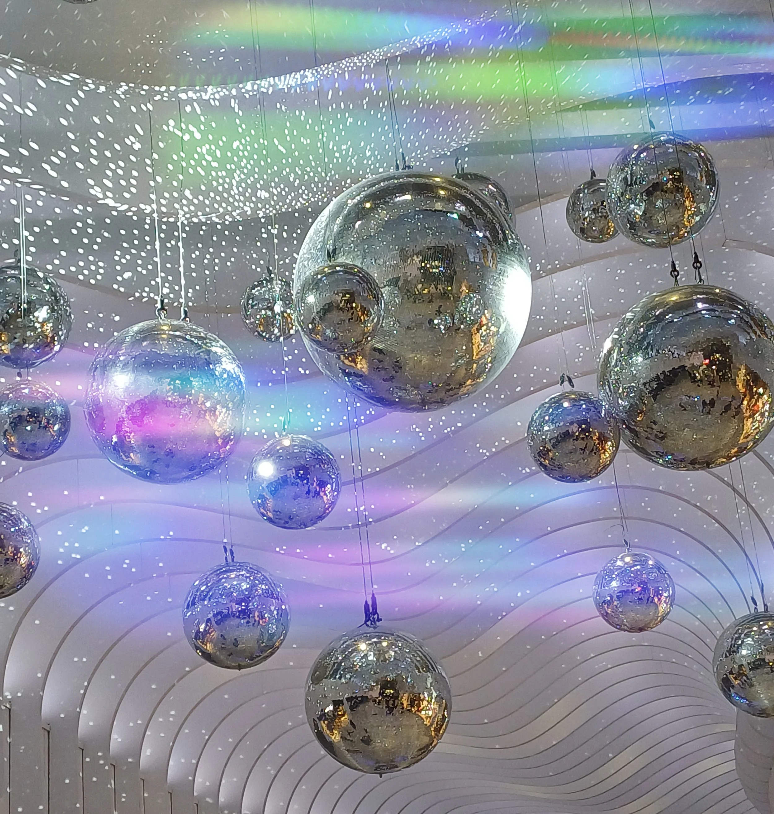 Multiple shiny silver spherical ornaments hanging from a ceiling, reflecting light and colors. The background features a white ceiling with circular light patterns and rainbow-colored reflections on the ornaments.