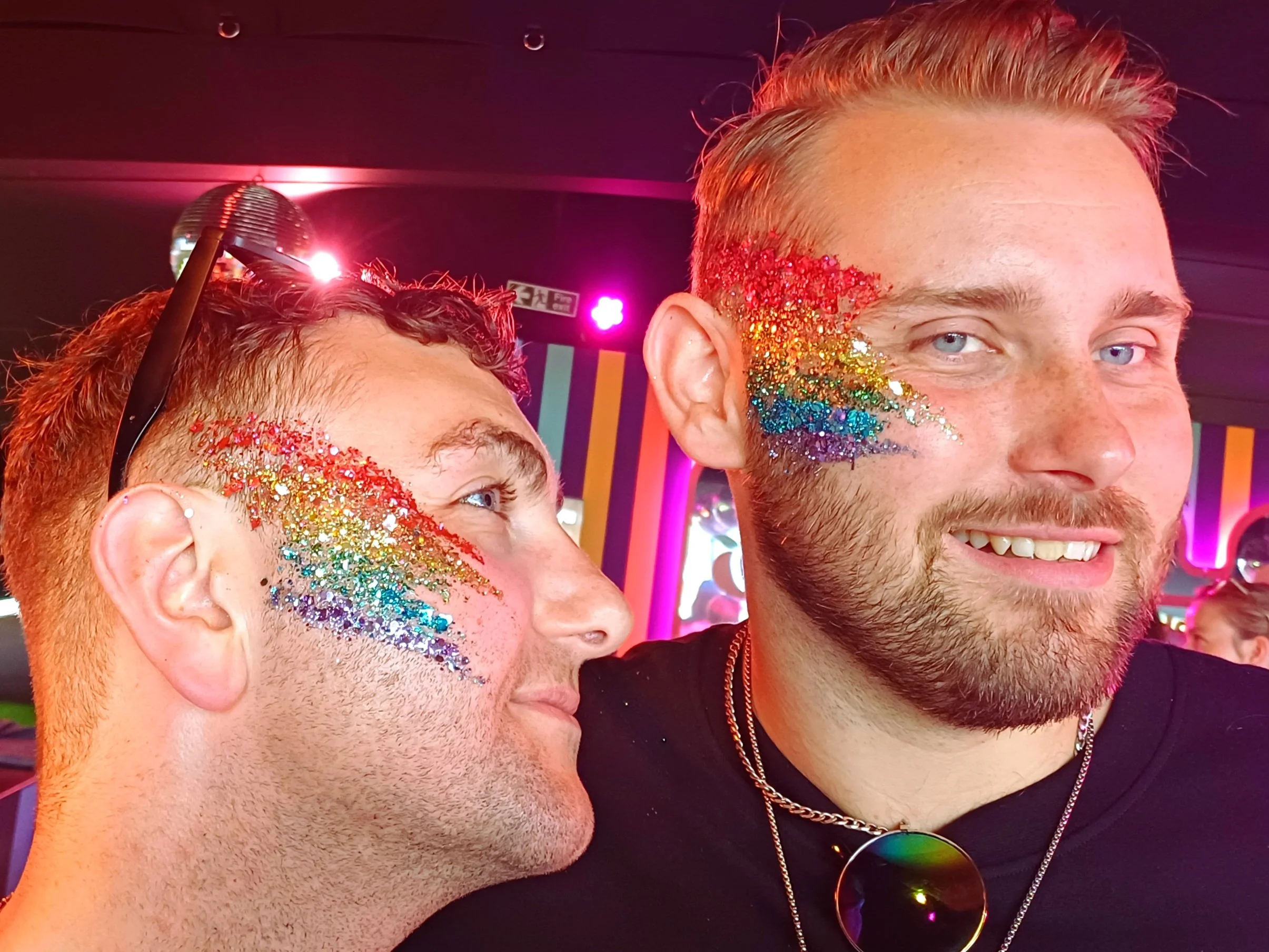 Two people with rainbow glitter face paint on their cheeks and near their eyes, close-up of their faces with a dark background and colorful lights.