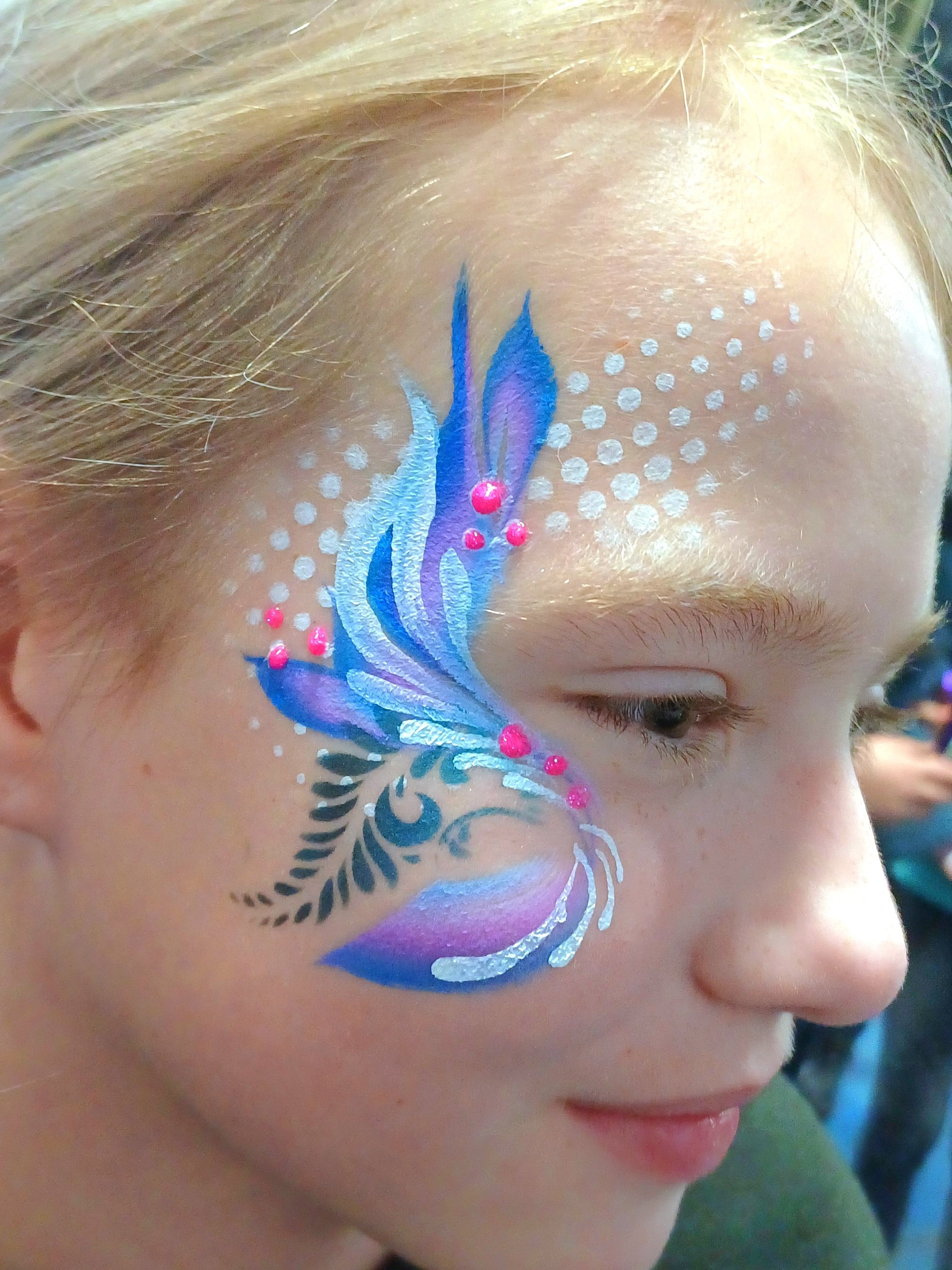 Close-up of a girl's face with a colorful festival face painting on her cheek, featuring shades of blue, purple, white, and pink, surrounded by small white dots.