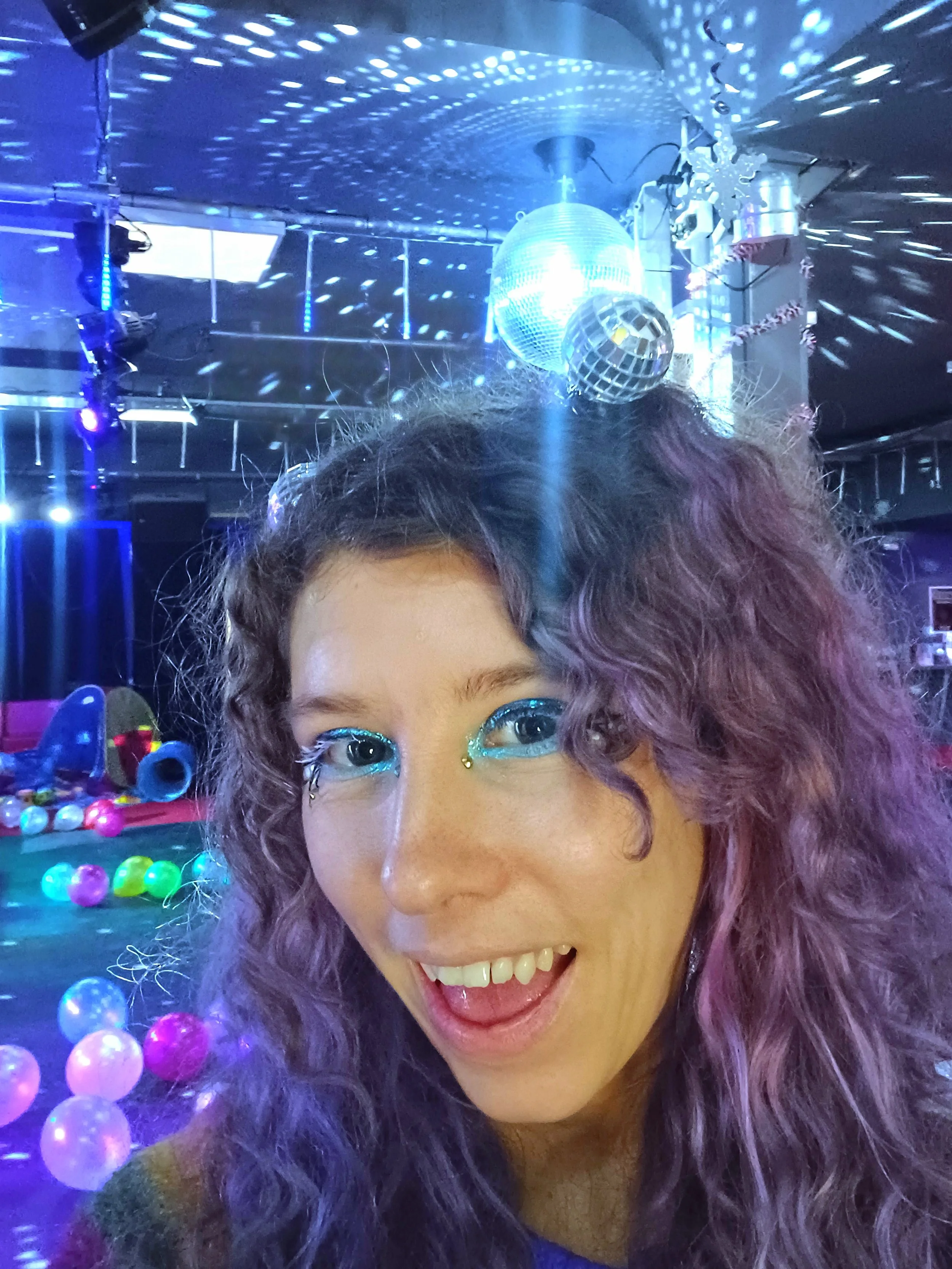 A woman with curly purple hair and bright makeup smiling at a party with disco balls and colorful balls in a dimly lit room.