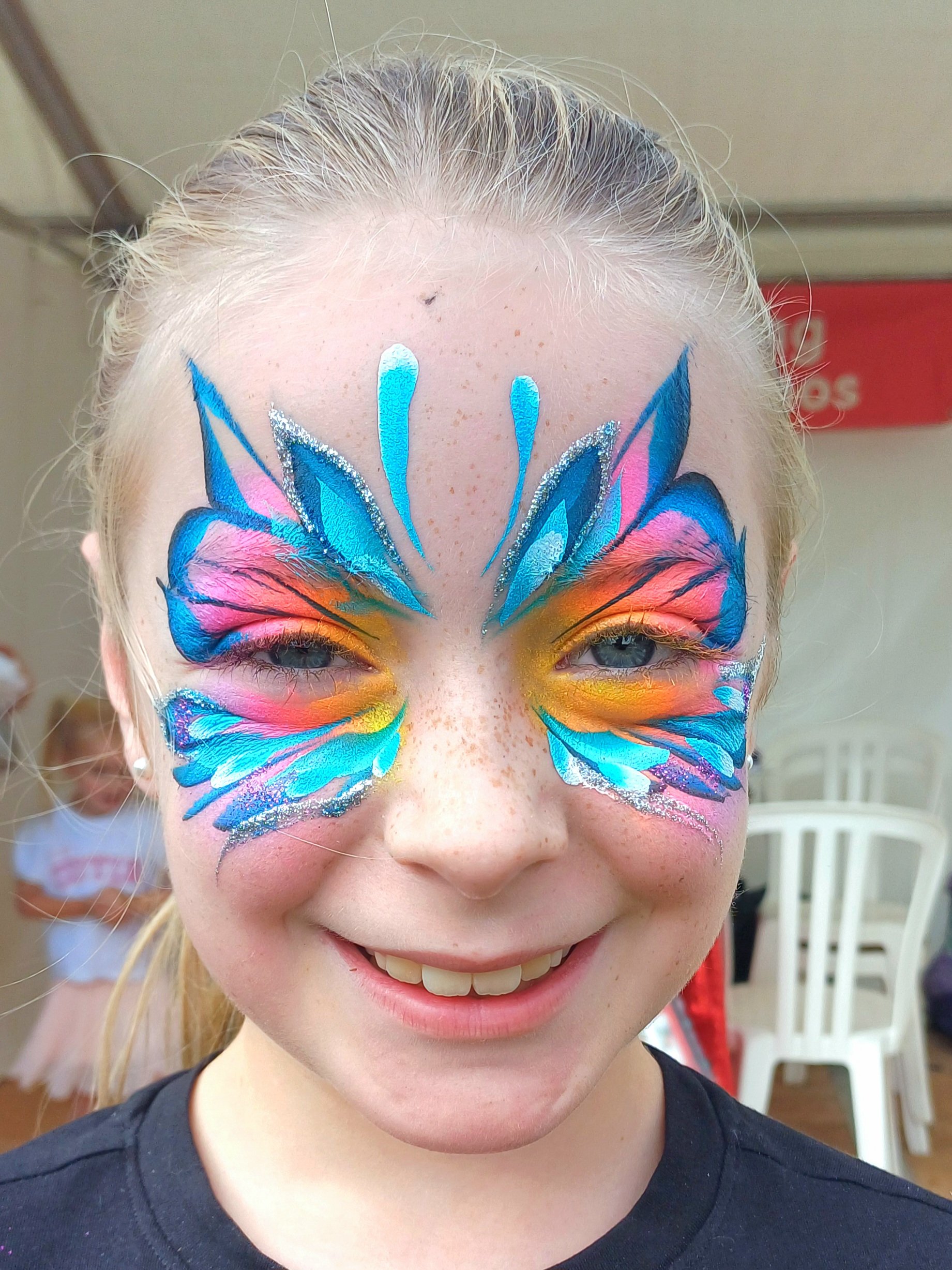 A woman with face painted as a colorful butterfly, smiling with visible teeth, and blonde hair tied back. In the background, there are white plastic chairs and a person in a pink shirt.