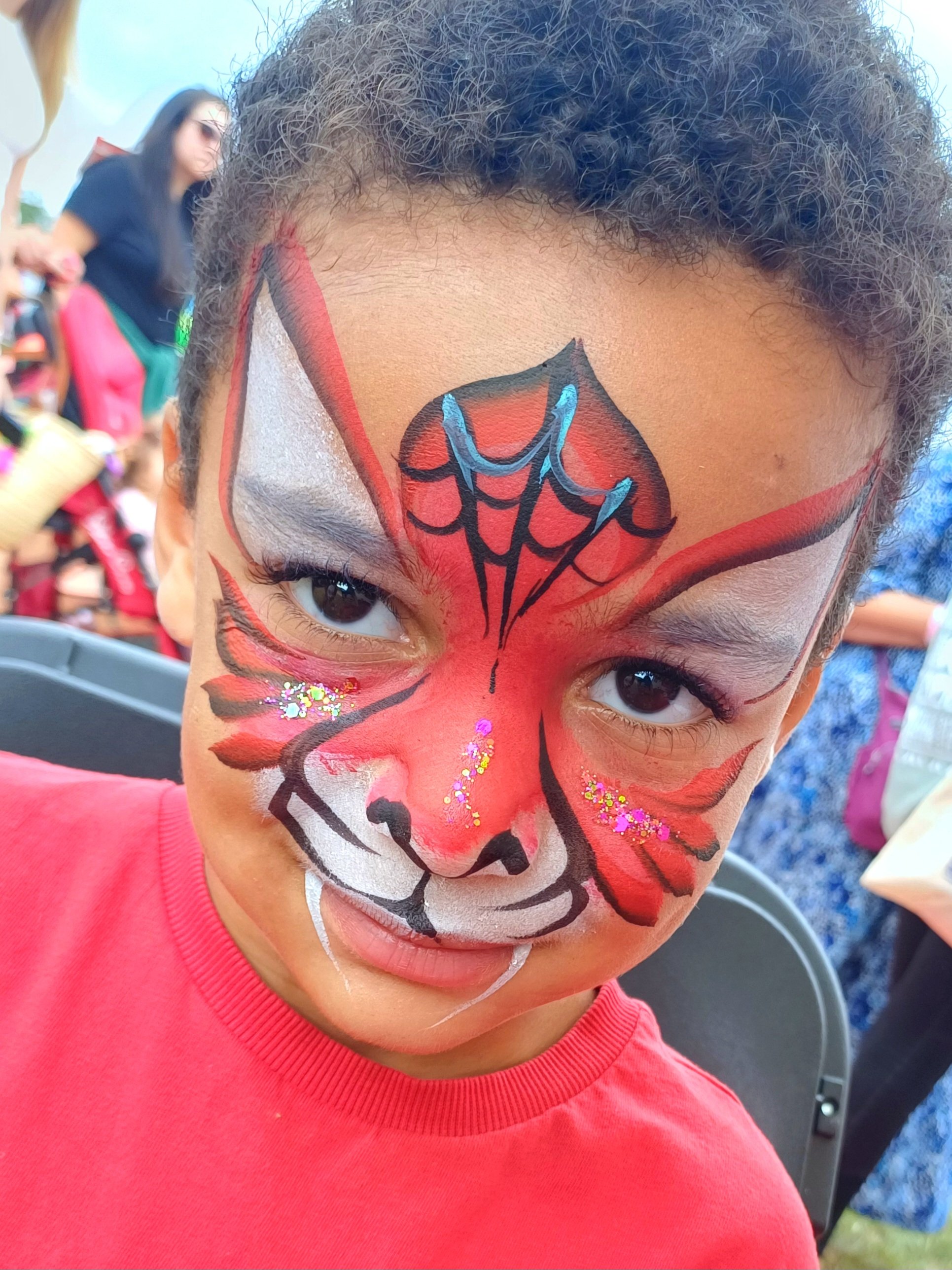 A person with spider-cat face paint wearing a bright pink shirt, at an outdoor event.