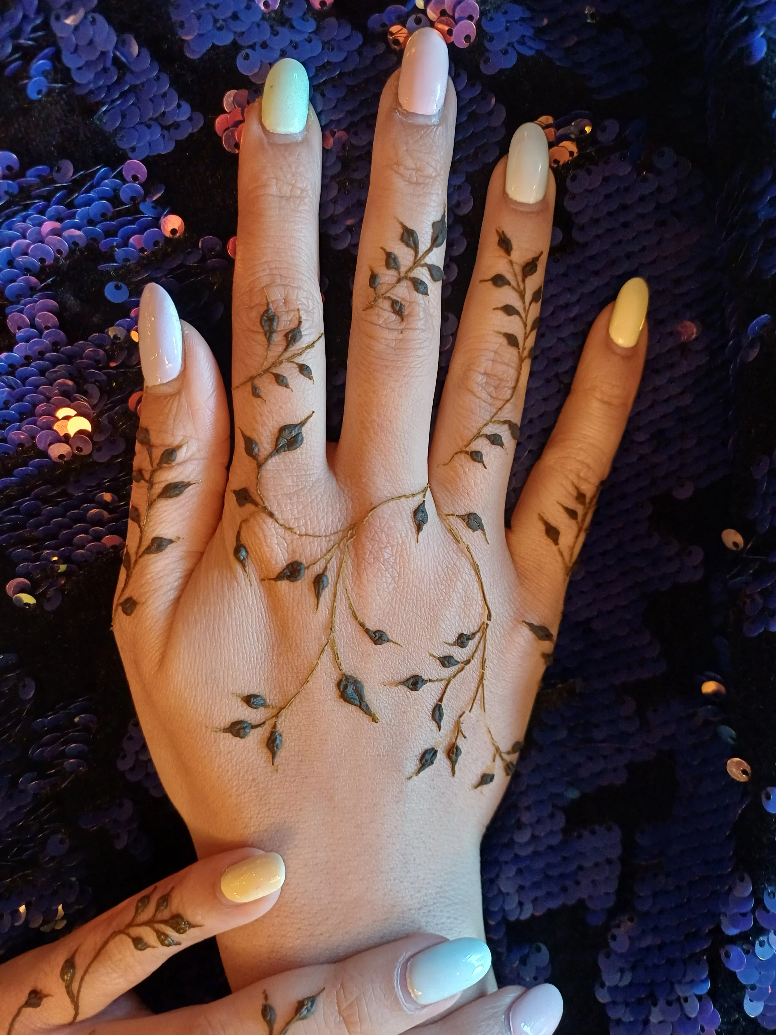 Close-up of a hand with intricate leaf-shaped tattoos on fingers and wrist, wearing light-colored and yellow nail polish, resting on a black fabric background with shiny purple sequins.