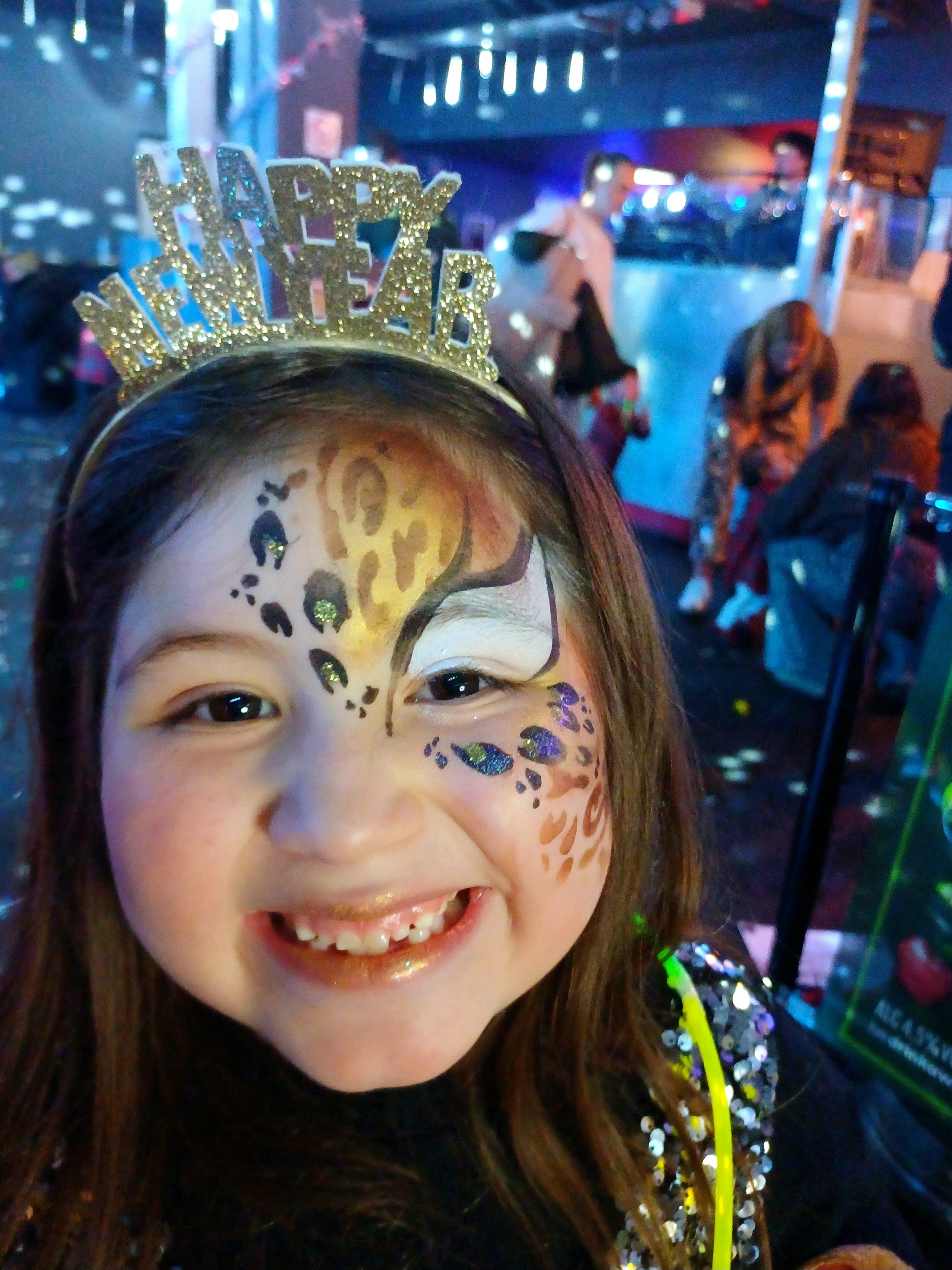 A young girl with face paint resembling a leopard, wearing a gold glittery 'Happy New Year' headband, smiling at a party with colorful lights and other people in the background.
