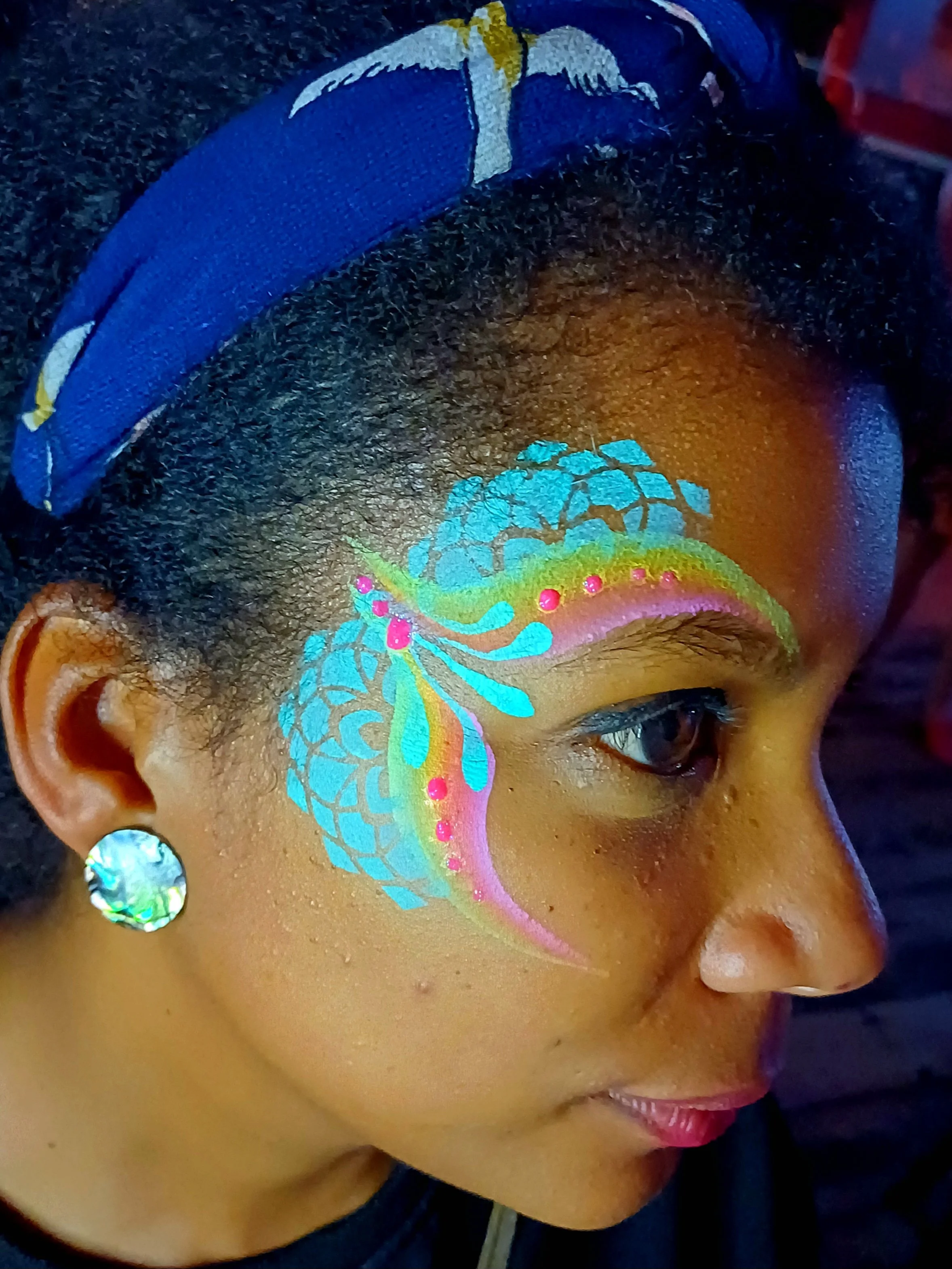 Close-up of a woman's face with vibrant, colorful face and eye makeup, including floral and intricate designs, along with a decorative earring and a dark background.