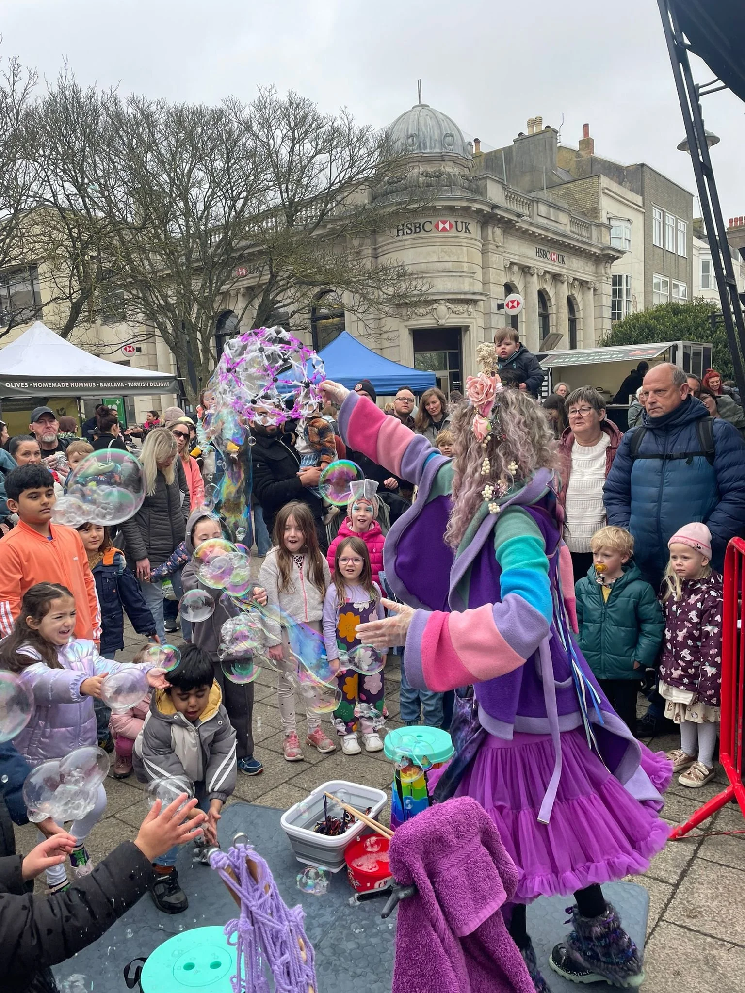 A street festival with children and adults gathered around a bubble performer dressed in vibrant, colorful costume. The performer is creating bubbles, entertaining the crowd on a cloudy day in front of an ornate building with trees and tents in the b