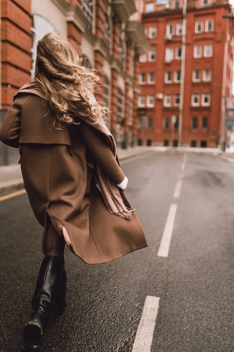 Woman in a brown coat walking along a quiet city street lined with red brick buildings.