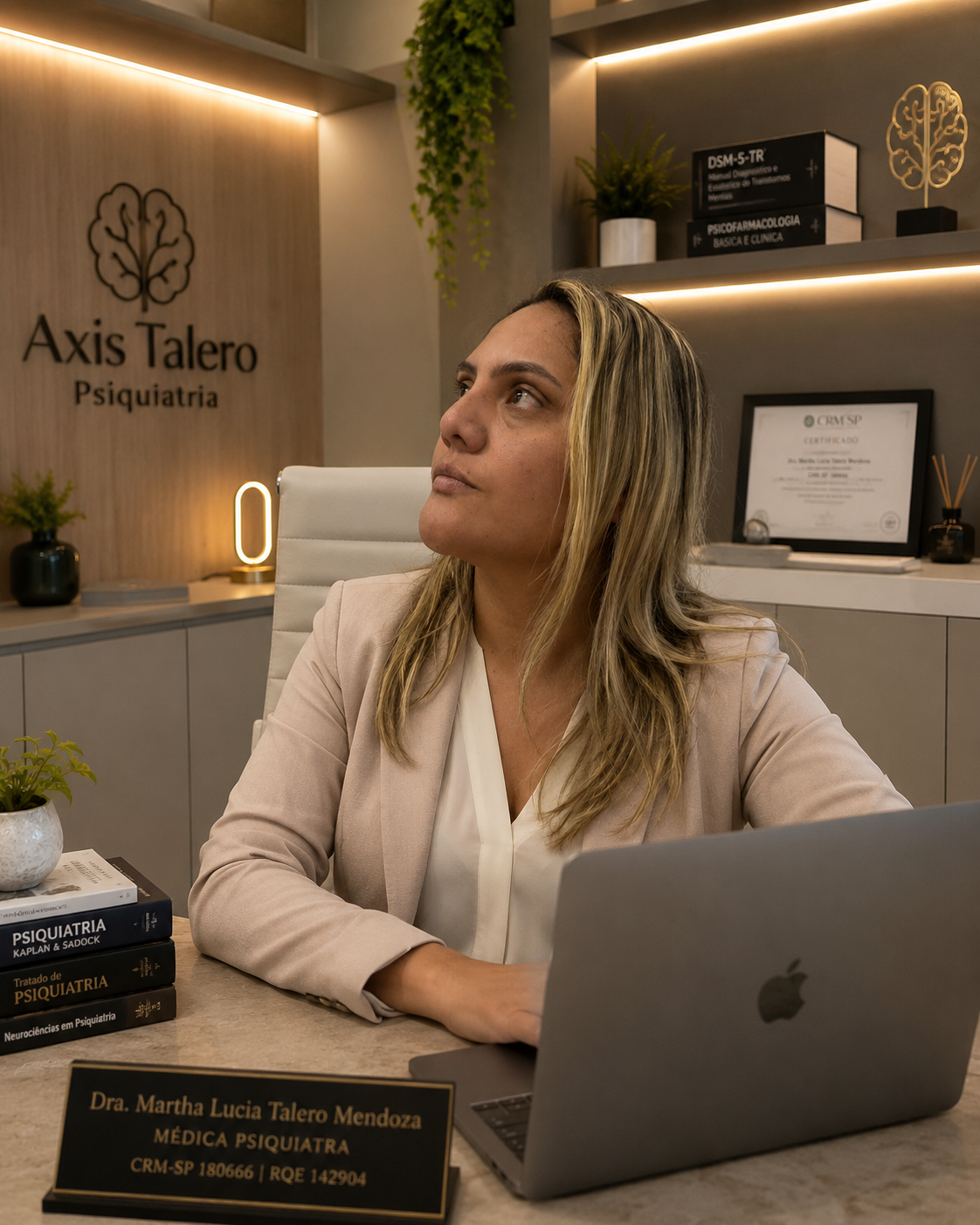 Dra. Martha Lucia Talero Mendoza, médica psiquiatra, em consultório com notebook, livros de psiquiatria e identificação da Axis Talero Psiquiatria.