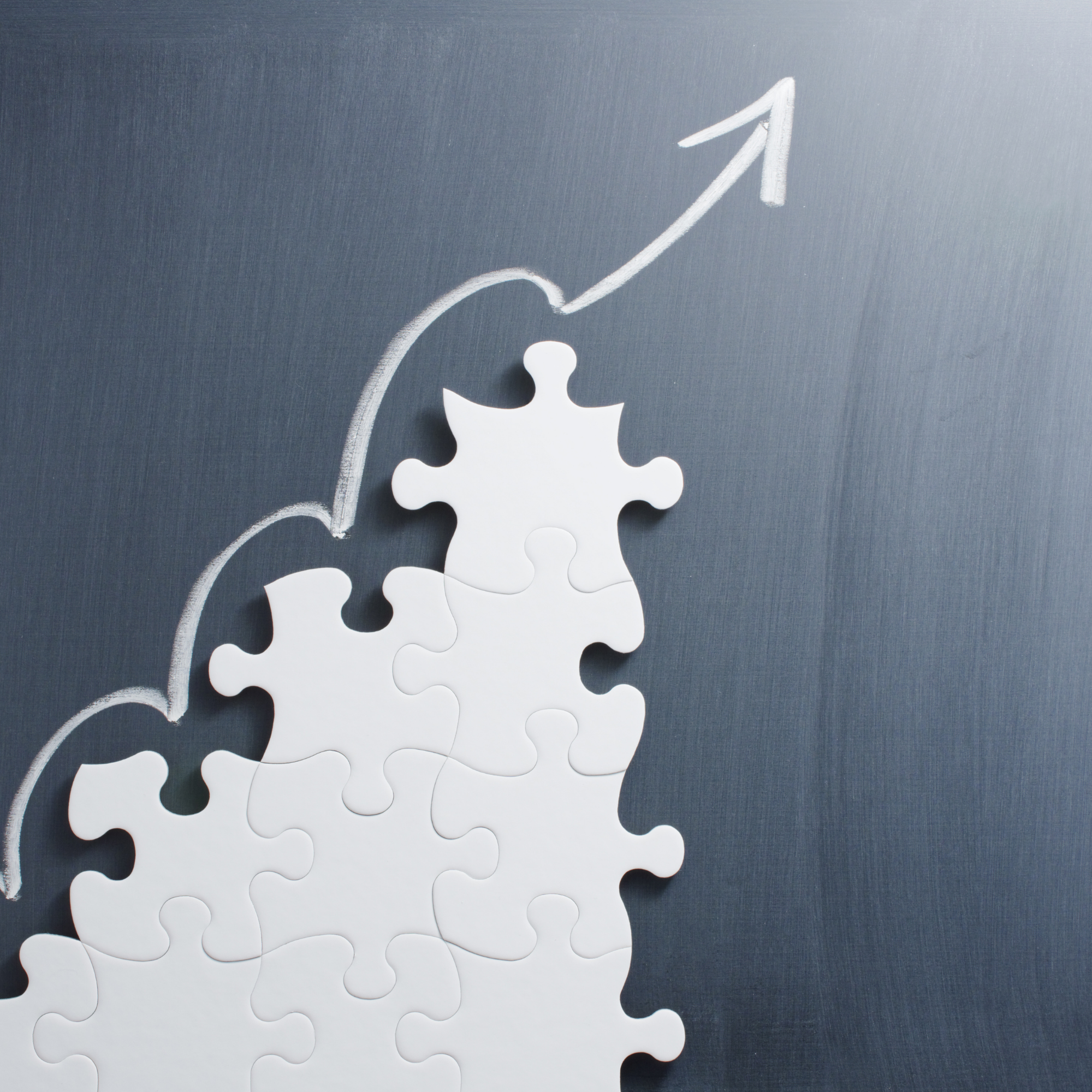 A piece of chalk was used to draw an upward wavy line with an arrow pointing upward on a gray chalkboard. There are white jigsaw puzzle pieces arranged increasing upward below the line.
