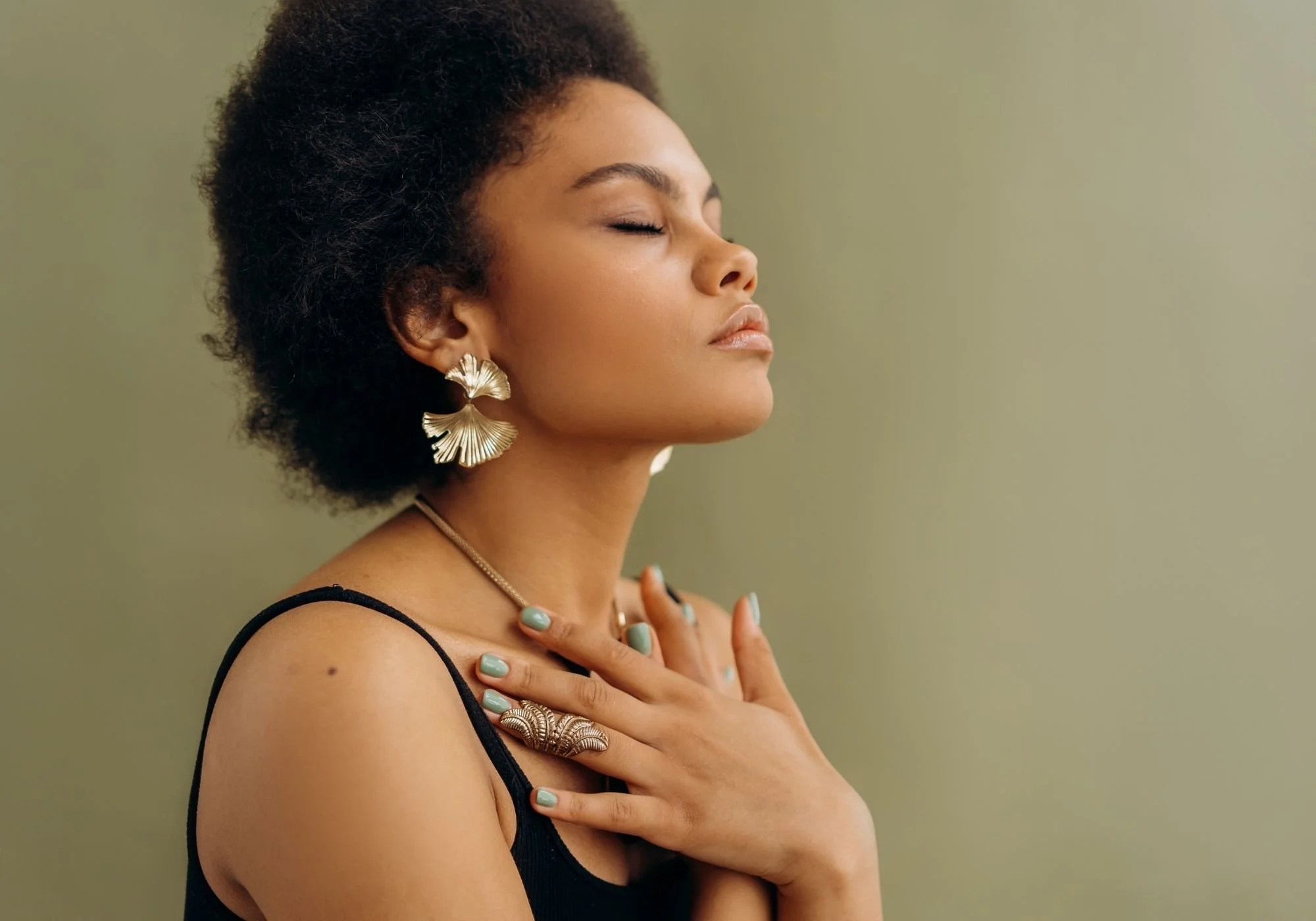 A woman with short curly hair and golden earrings with a leaf design, her eyes closed, wearing a black spaghetti strap top, resting her hand on her chest with mint green painted nails, against a plain green background.