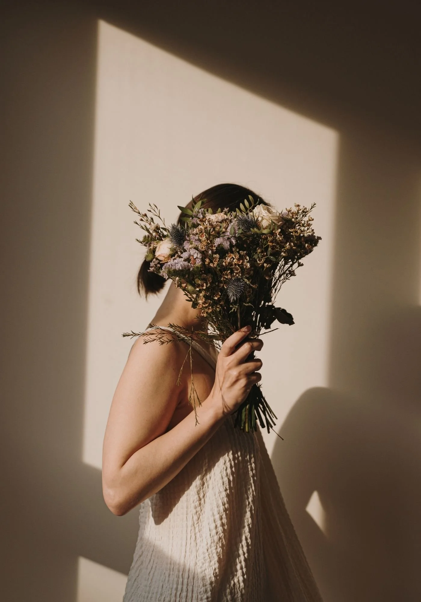Woman with long dark hair holding a large bouquet of dried flowers up in front of her face, standing in warm sunlight and shadow.