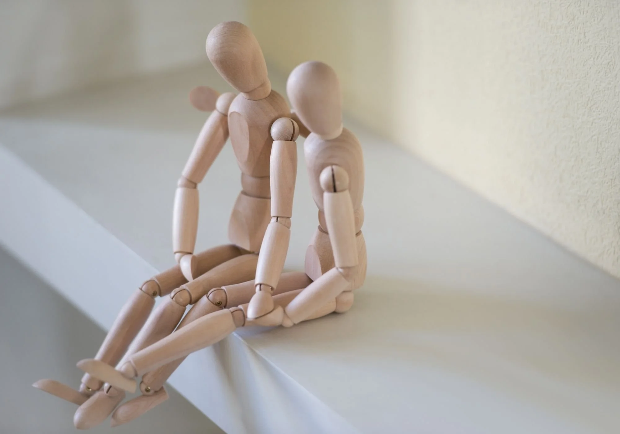 2 wooden mannequins sitting on a white surface near a yellow wall, one has it's head down and the other has its hand around it's shoulders.