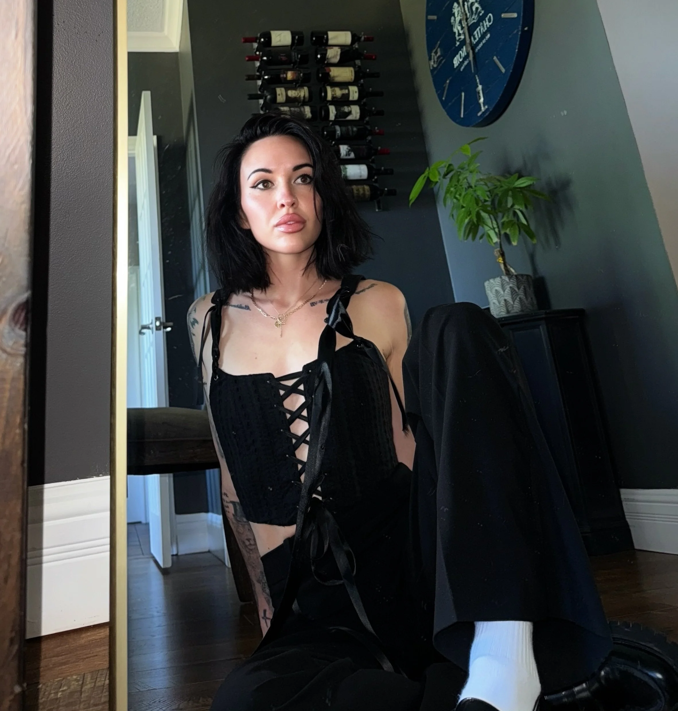 A woman with black hair and tattoos on her shoulder, wearing black clothing, is looking into a mirror in a room with a wall of wine bottles, a soccer-themed clock, and a potted plant.