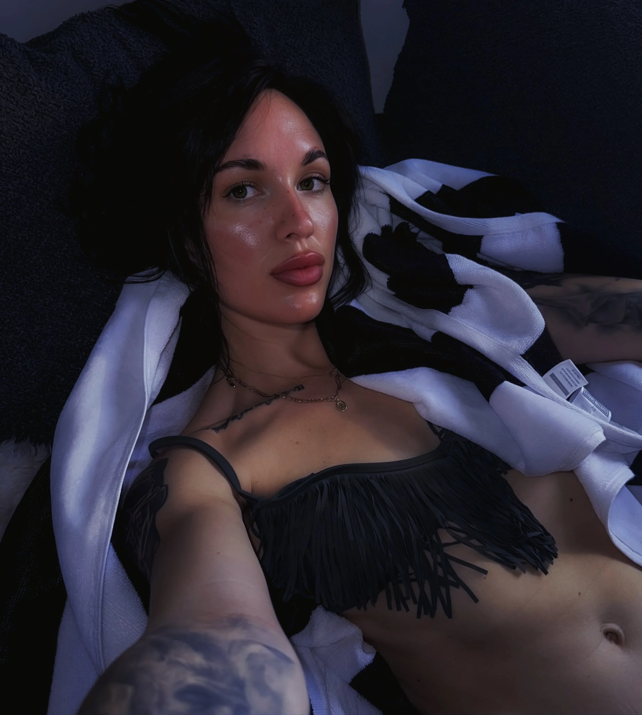 A woman lying down on a black surface, wearing a black and white striped shirt with fringe details and a necklace, with dark hair and makeup, looking at the camera.