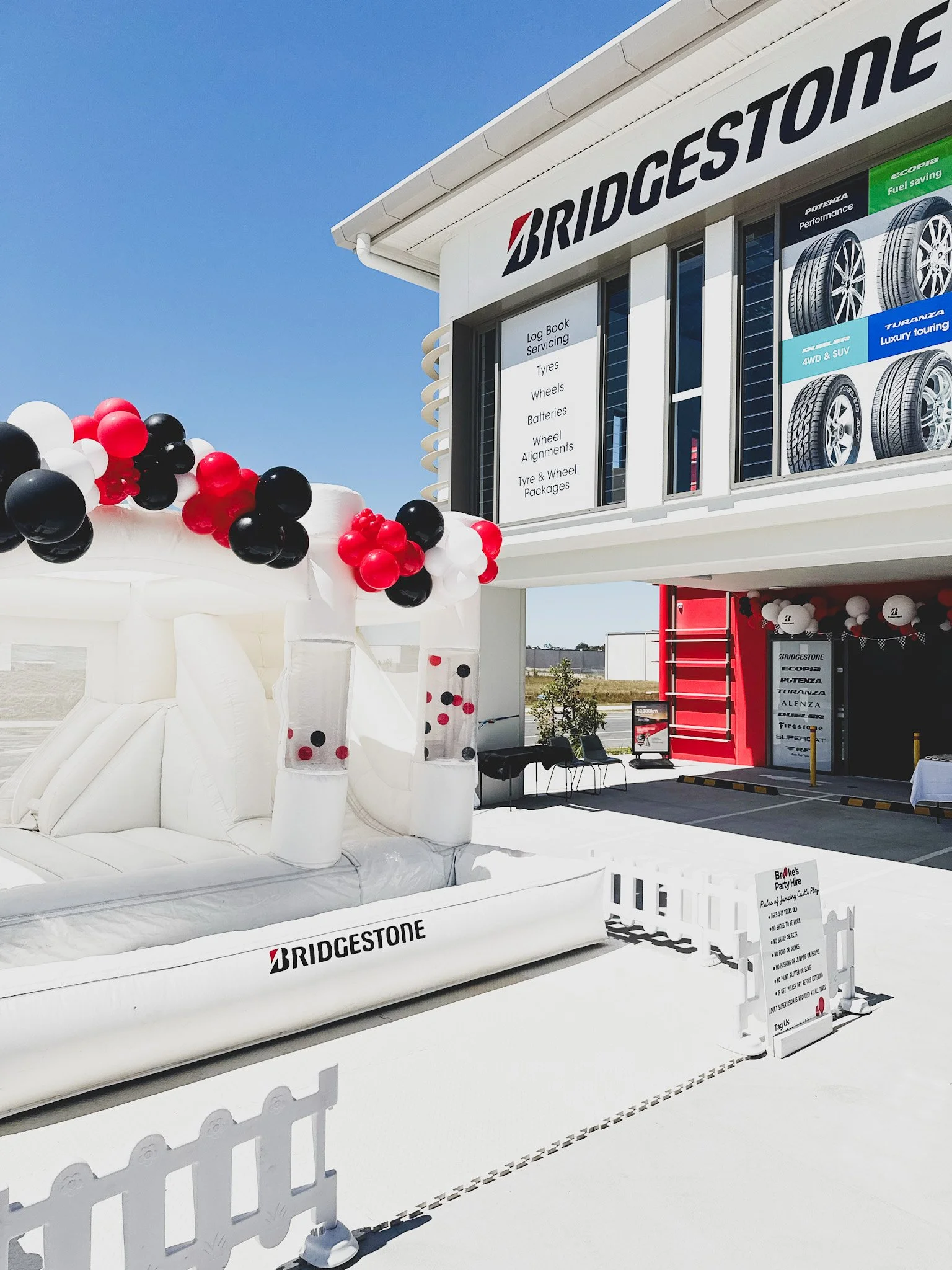 Bridgestone Select tyre store with a white inflatable bounce house decorated with black, white, and red balloons, and a fenced-off area with a sign showing store policies, during a bright, sunny day.