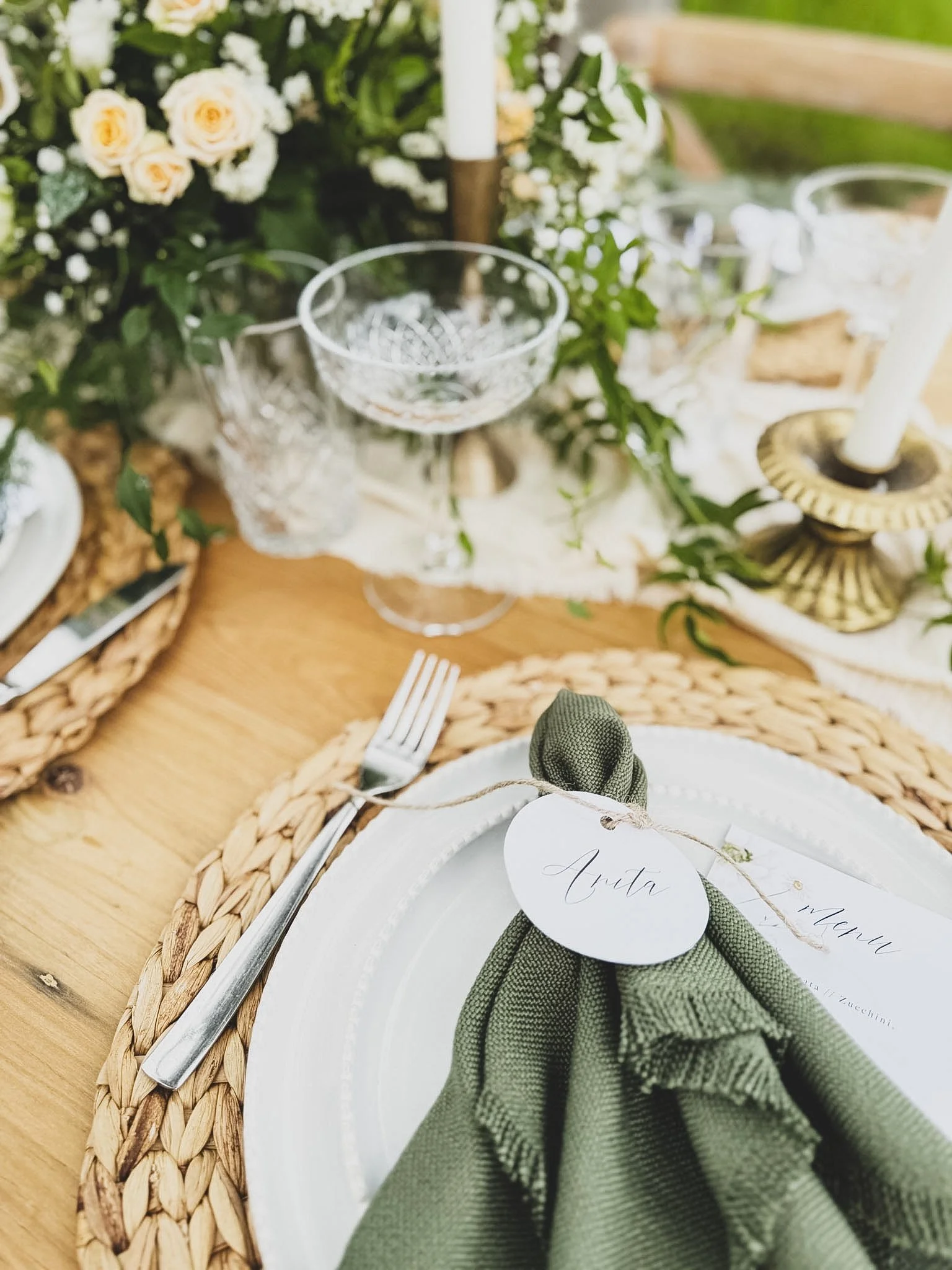 Close-up of a table setting with a woven placemat, greenery, glassware, and a white plate with a folded green cloth napkin with a name tag reading 'Anita'.