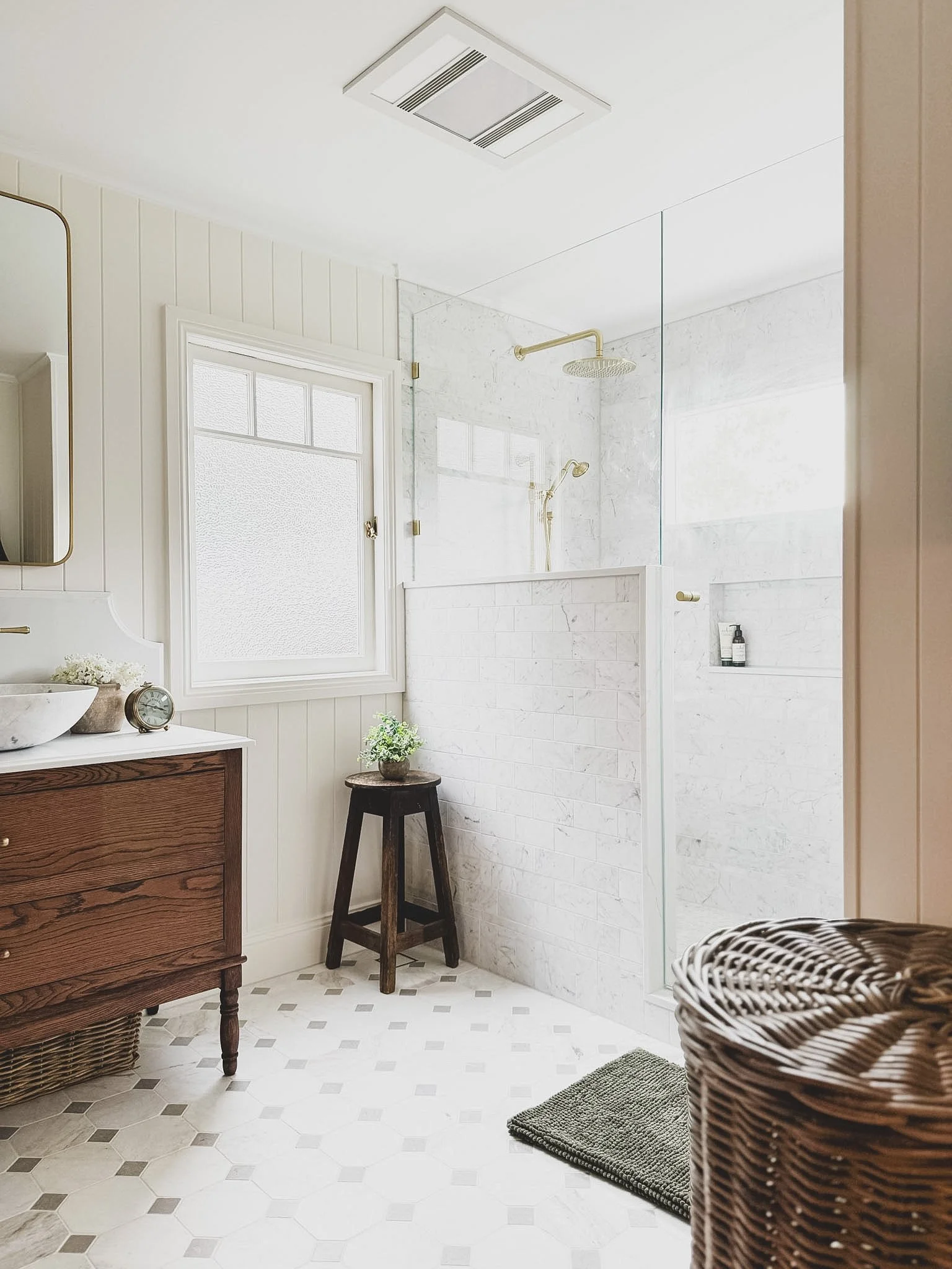 Bright, modern bathroom with white tiled shower area, glass partition, and brass fixtures. Features a wooden vanity with a vessel sink, a mirror, a small clock, a potted plant, and a textured gray rug. Wicker basket in the foreground.