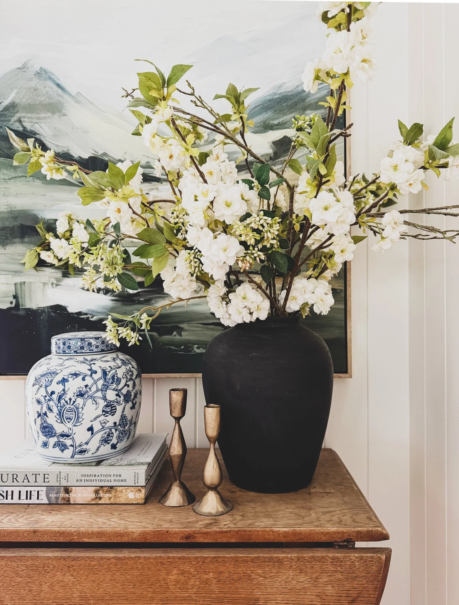A decorative arrangement of white flowers and green leaves in a black vase, placed on a wooden surface with a painting and blue and white patterned vase in the background.