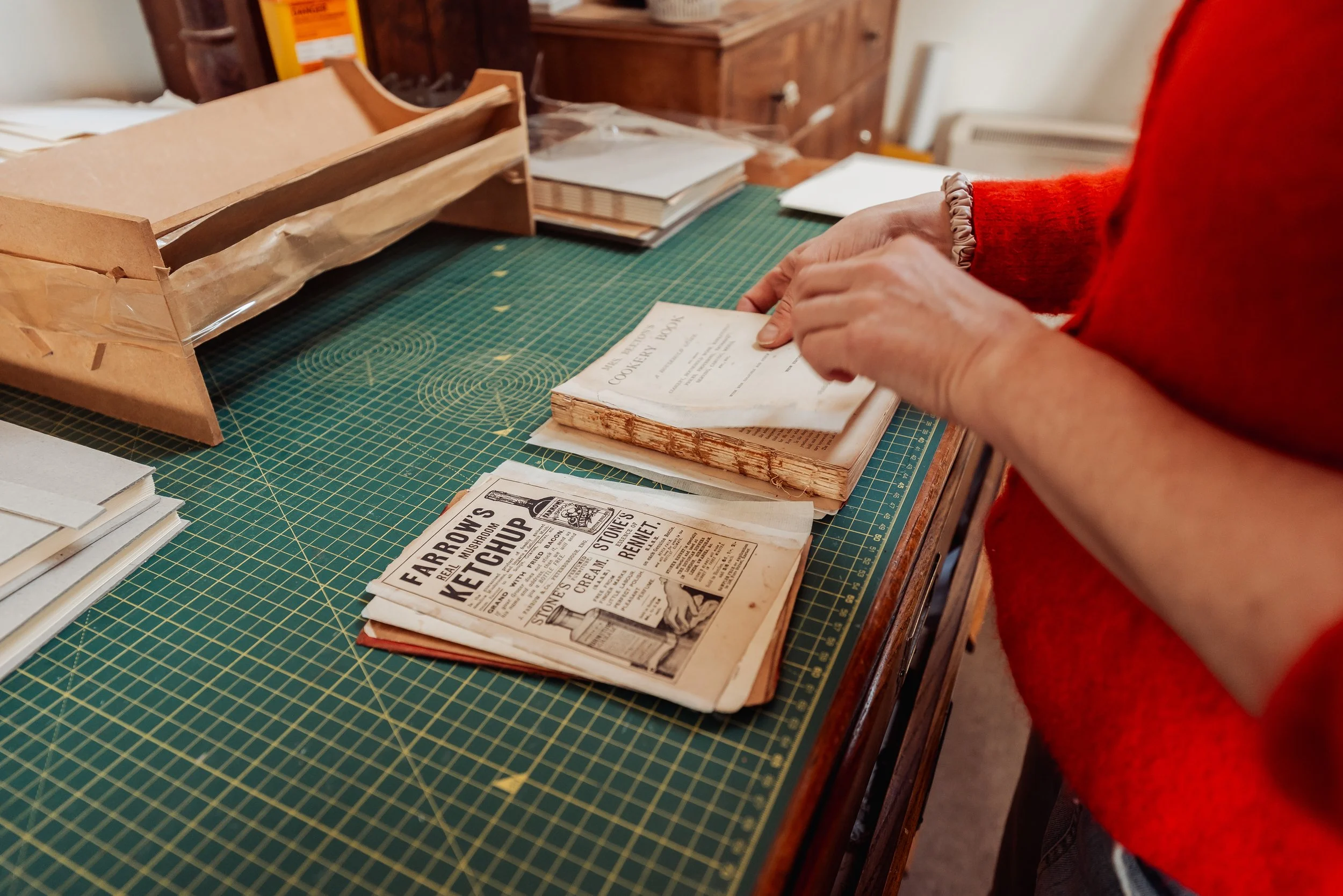 Person in a red sweater organizing vintage newspapers and documents on a green cutting mat in a workspace.