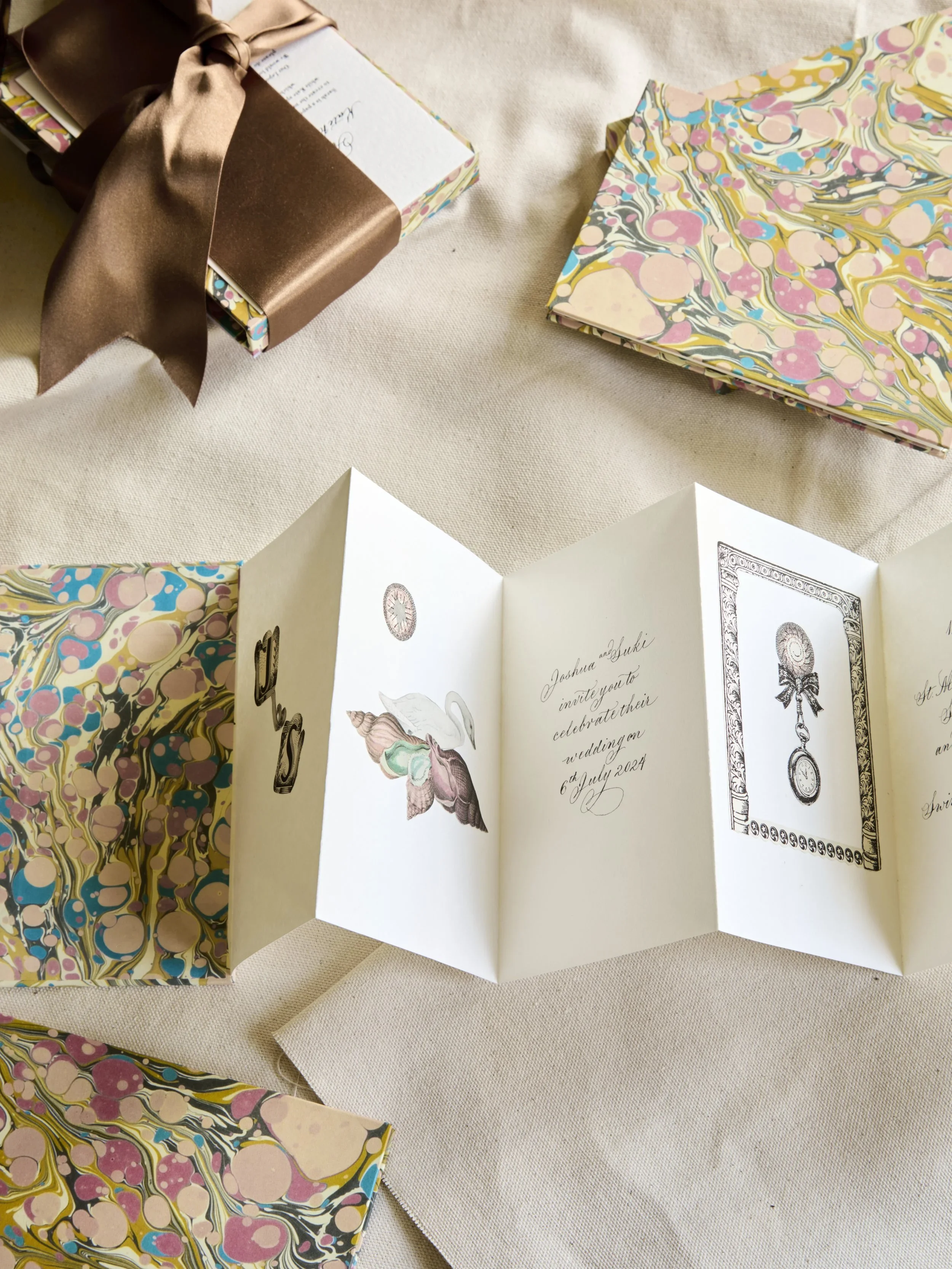 A pop-up greeting card with floral illustrations, and a wedding invitation inside. Also visible are decorated gift boxes on a beige fabric surface, one wrapped with a bronze satin ribbon.