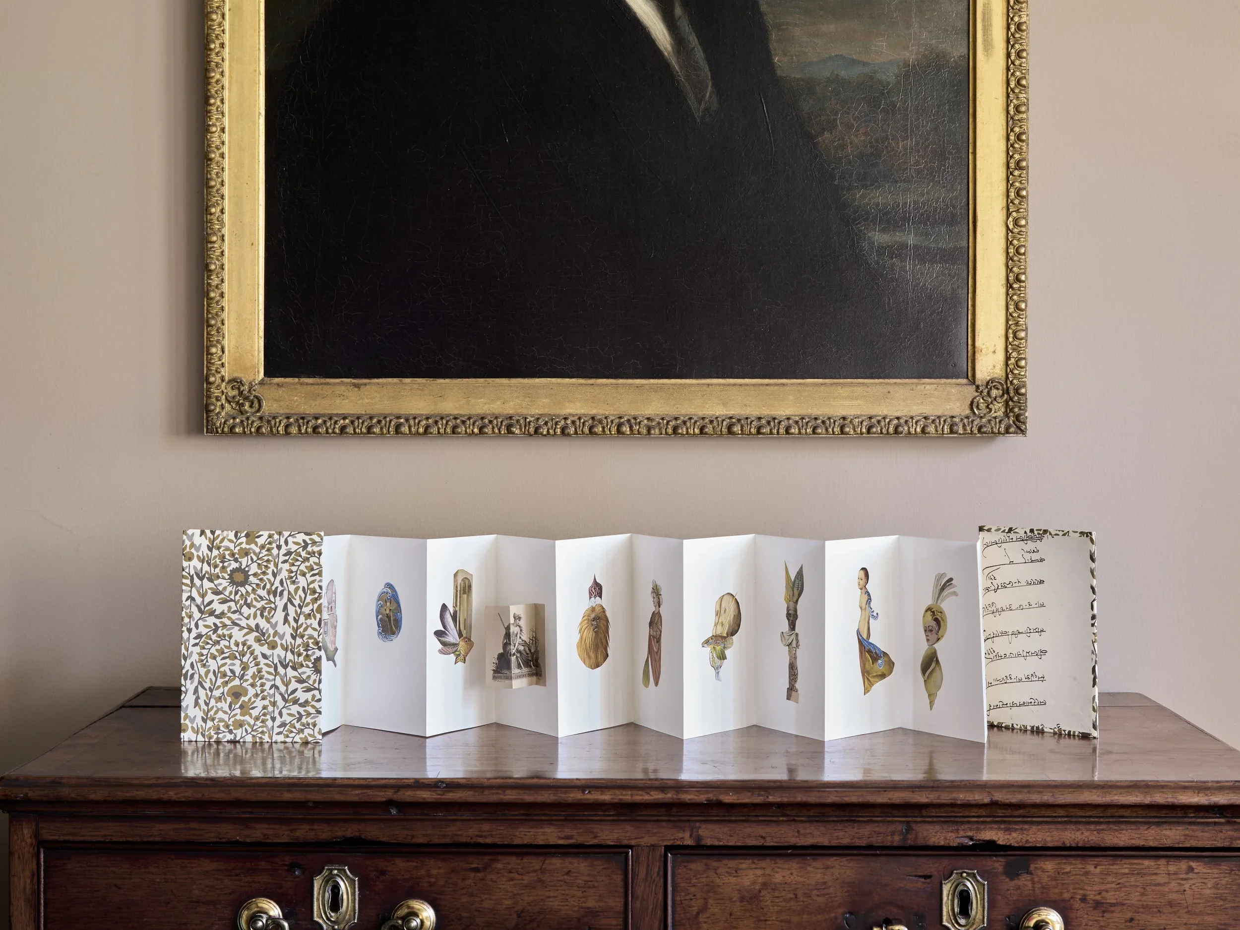 A large framed painting hangs on a beige wall. Below it is a wooden table with a display of paper cutouts of birds and other objects, arranged in a zigzag pattern, with a decorative sheet of paper with handwritten notes to the right.