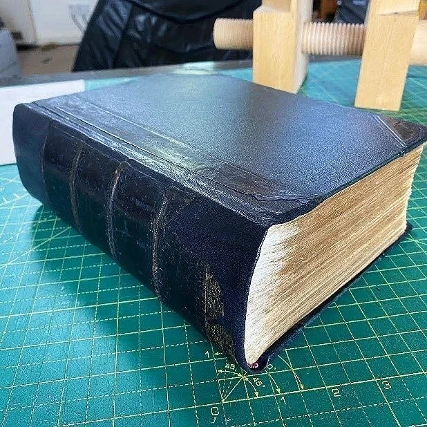 A large, old black leather-bound book with gold-edged pages resting on a green cutting mat.
