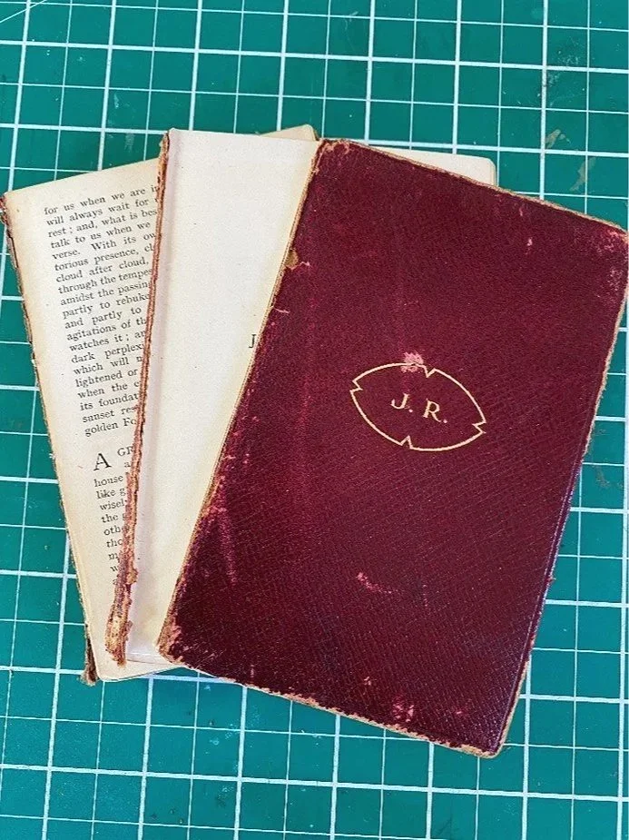 Three old worn books stacked on a green cutting mat, with the top book having a dark red cover and the initials 'J. R.' in gold on the front.