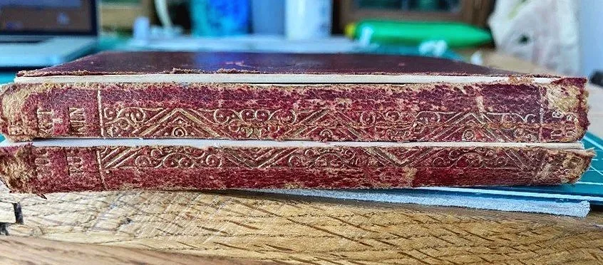 Close-up of an old, worn book with a red cover and embossed gold decorative patterns on the spine, resting on a wooden surface.