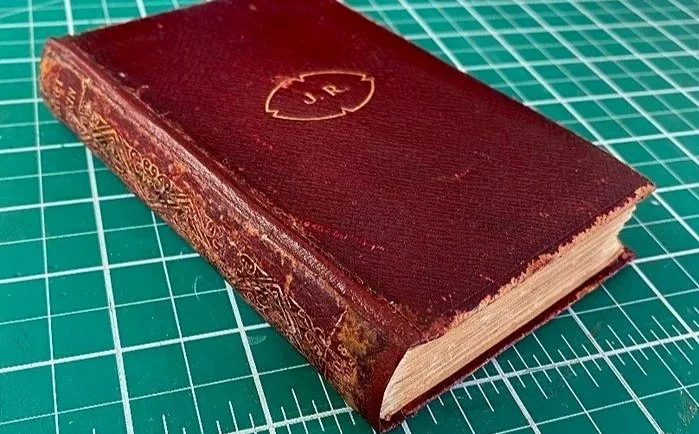 Close-up of an old, worn red hardcover book with gold embossed initials "J R" on the cover and intricate designs on the spine, placed on a green cutting mat with measurement markings.
