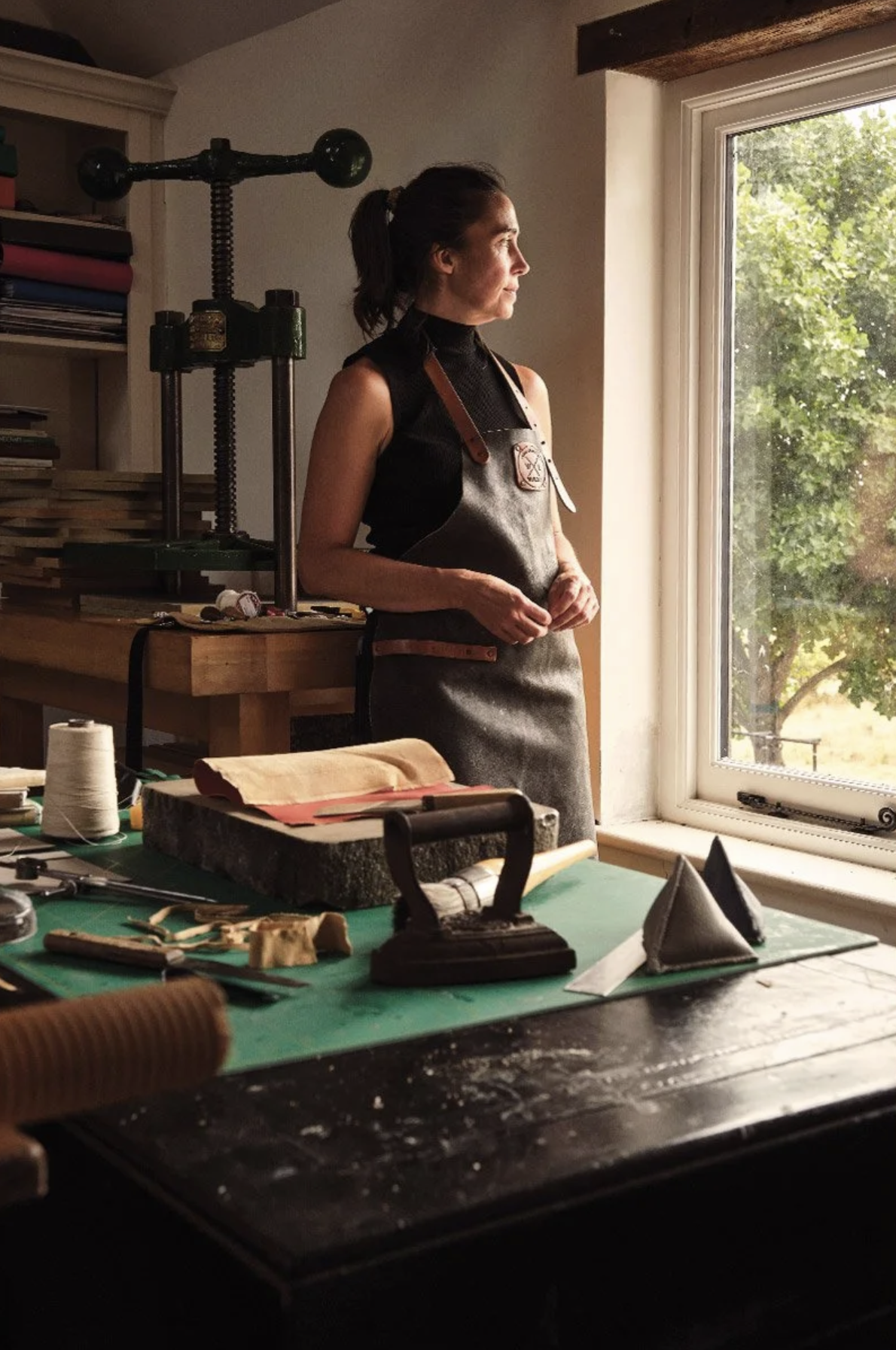 A woman in a black sleeveless top and gray apron standing by a window, in a workspace with tools and materials on a table, looking outside.