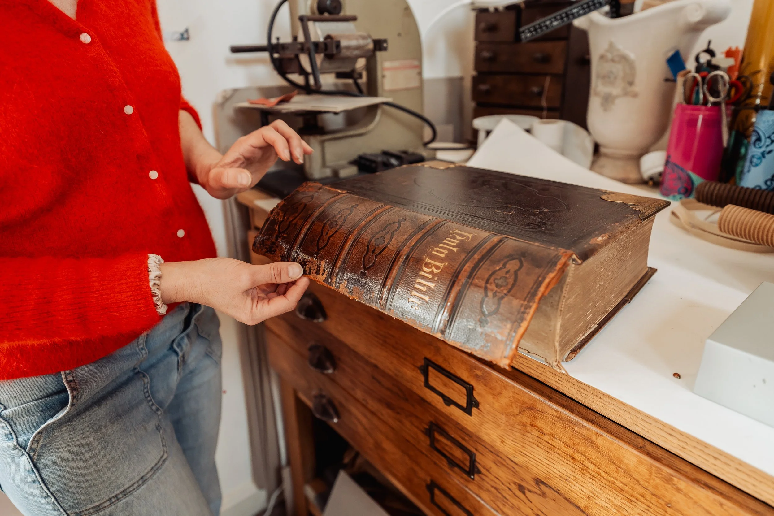 Person in a red sweater examining an old leather-bound book at a workspace with vintage equipment and various supplies.