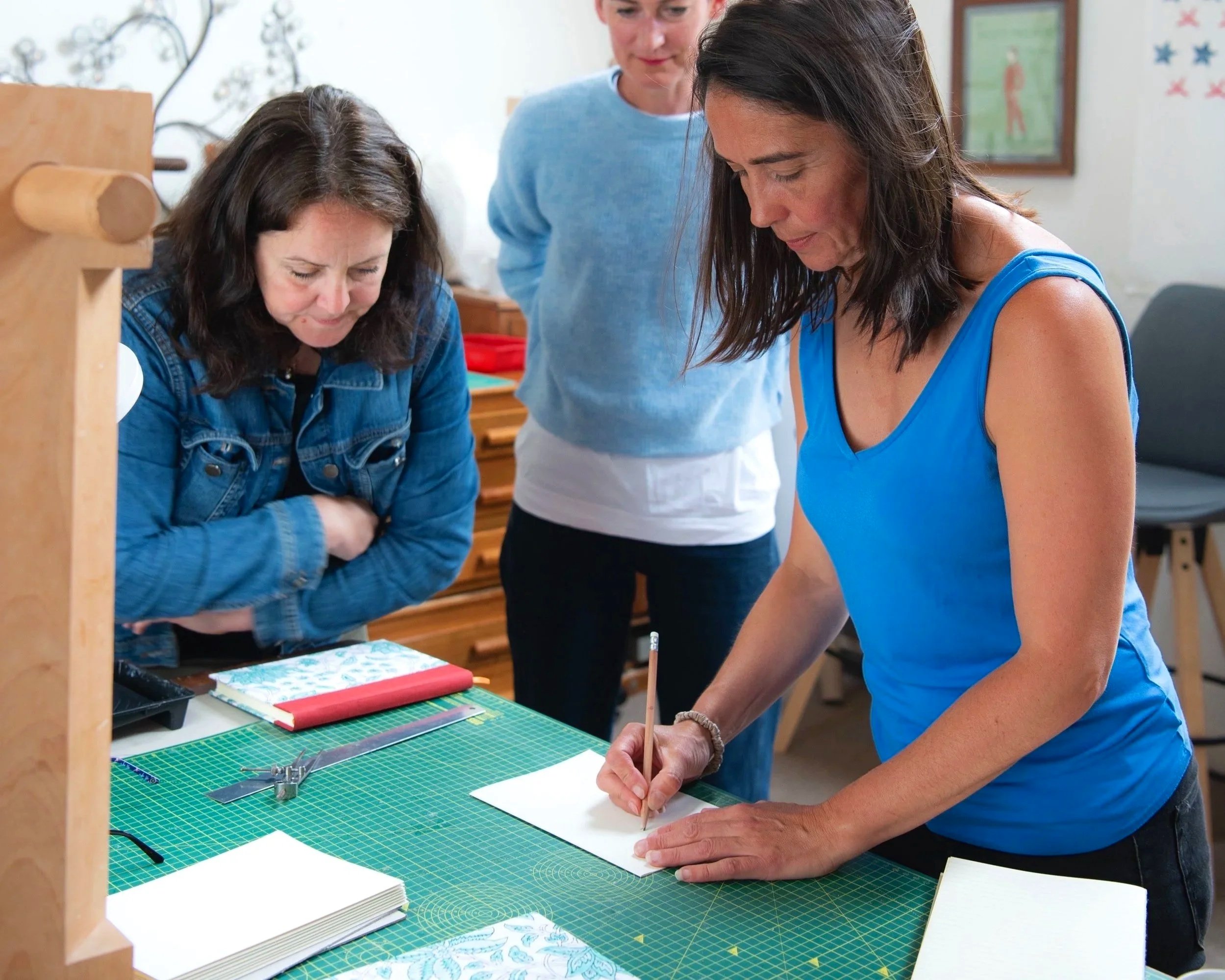 Three women around a green cutting mat working on a crafting project, with notebooks and tools on the table.