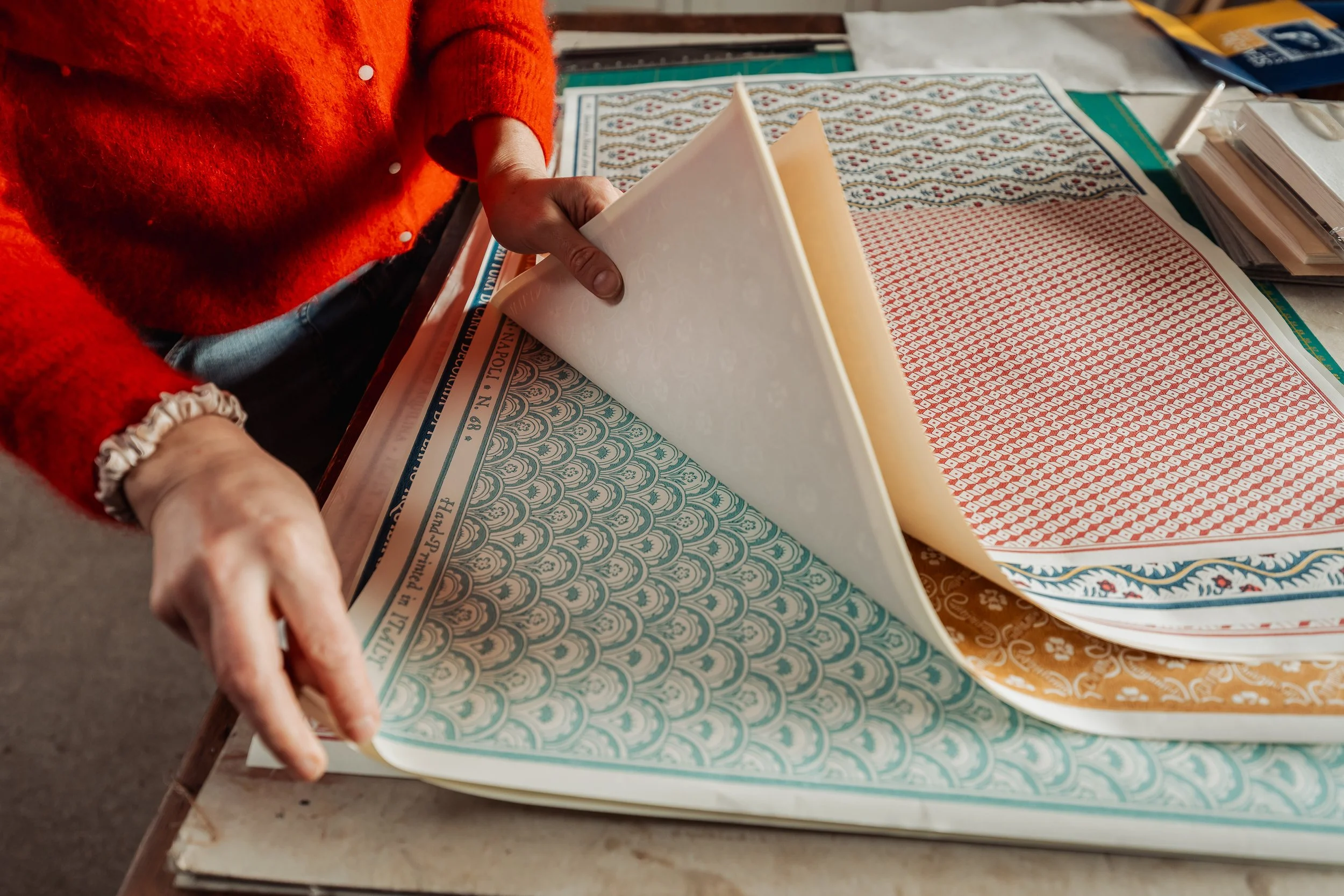 A person in a red sweater is flipping through patterned paper sheets on a cluttered table.