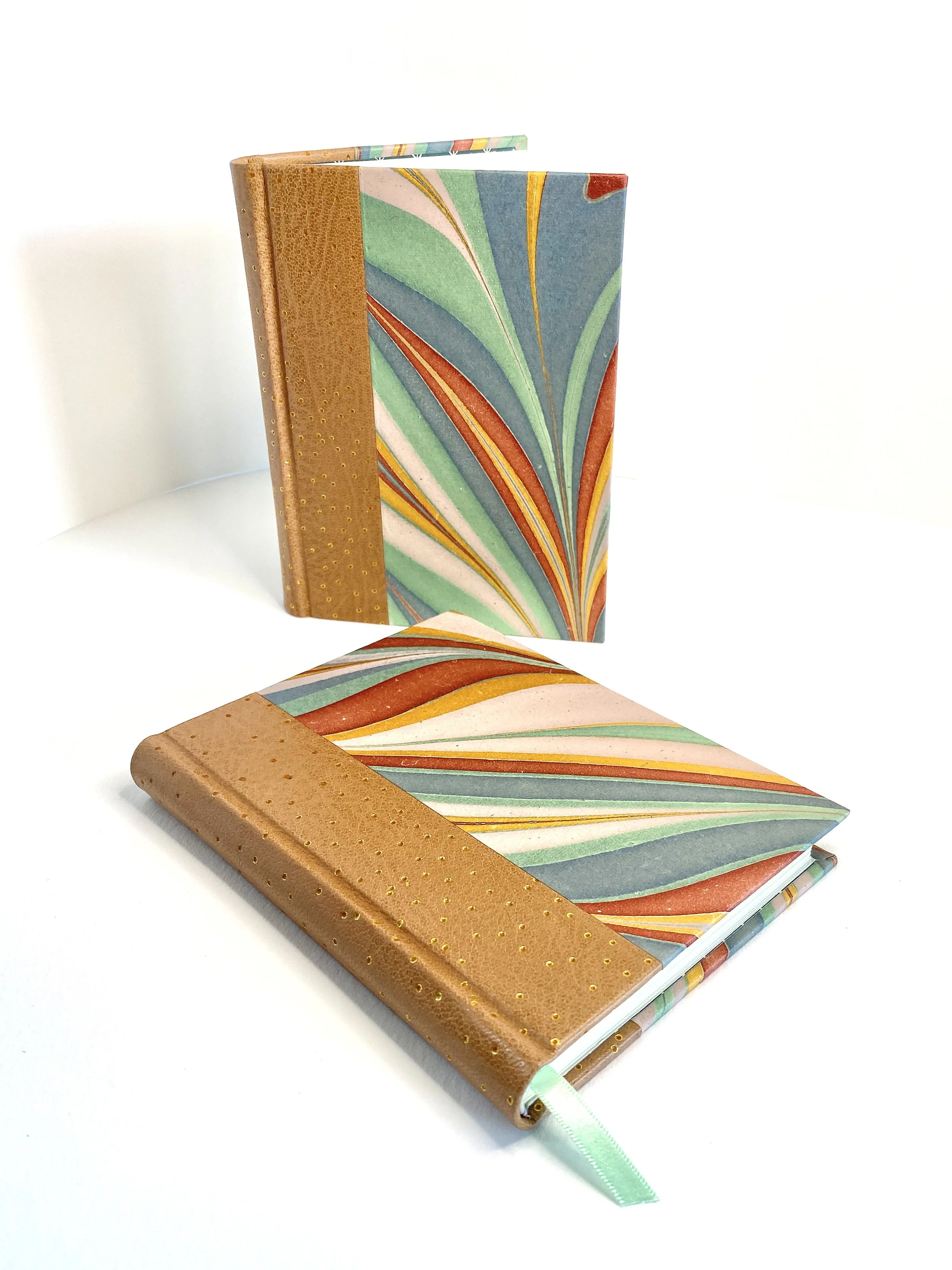 Two notebooks with decorative covers; one standing upright, the other lying flat. The standing notebook has a multicolored marbled cover, and the flat notebook has a light brown textured spine. A green ribbon marker extends from the closed notebook.