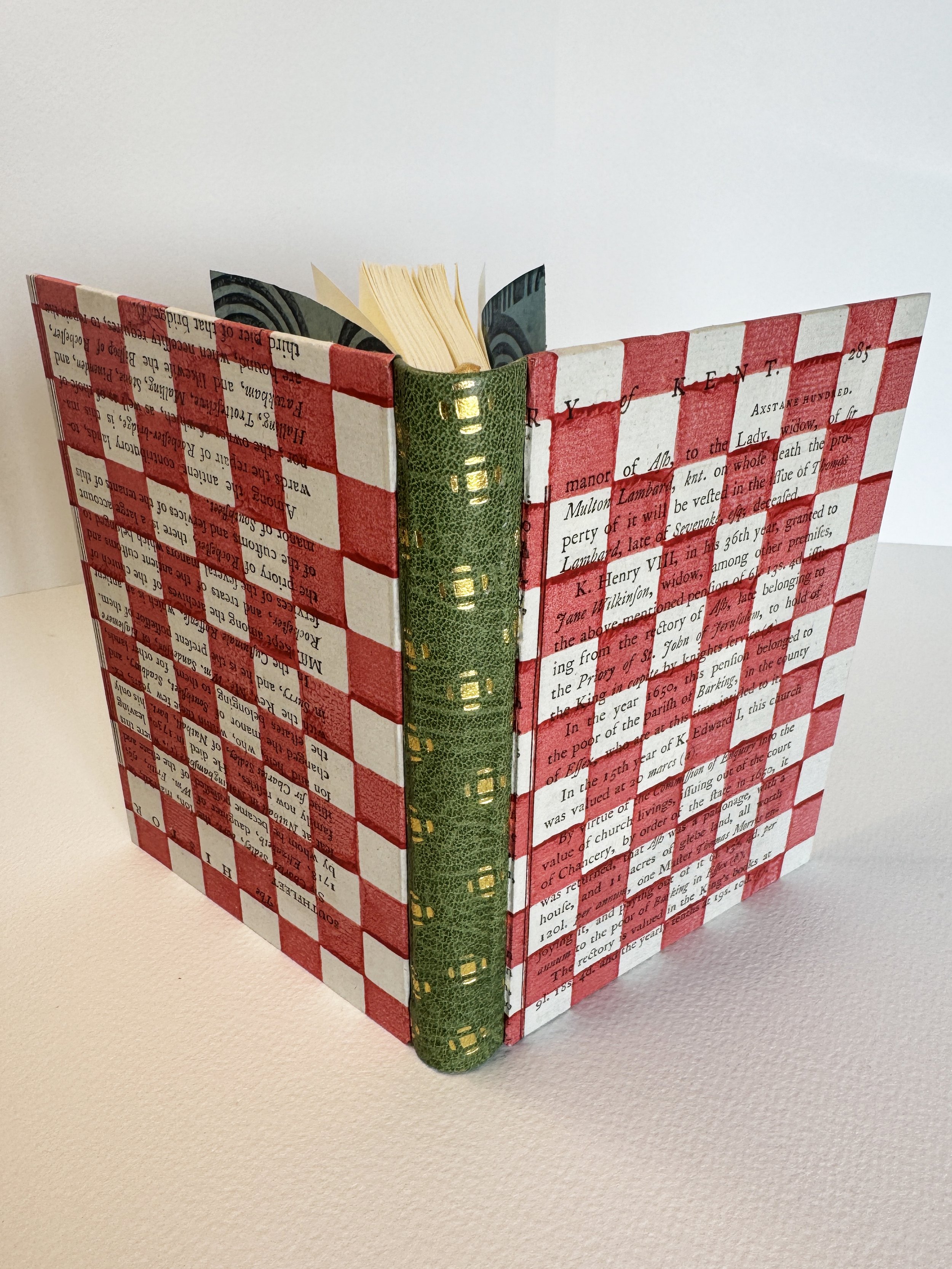 A decorative book with a red and white checkered cover, black text, and a green spine with gold accents, opened and standing upright on a white surface.