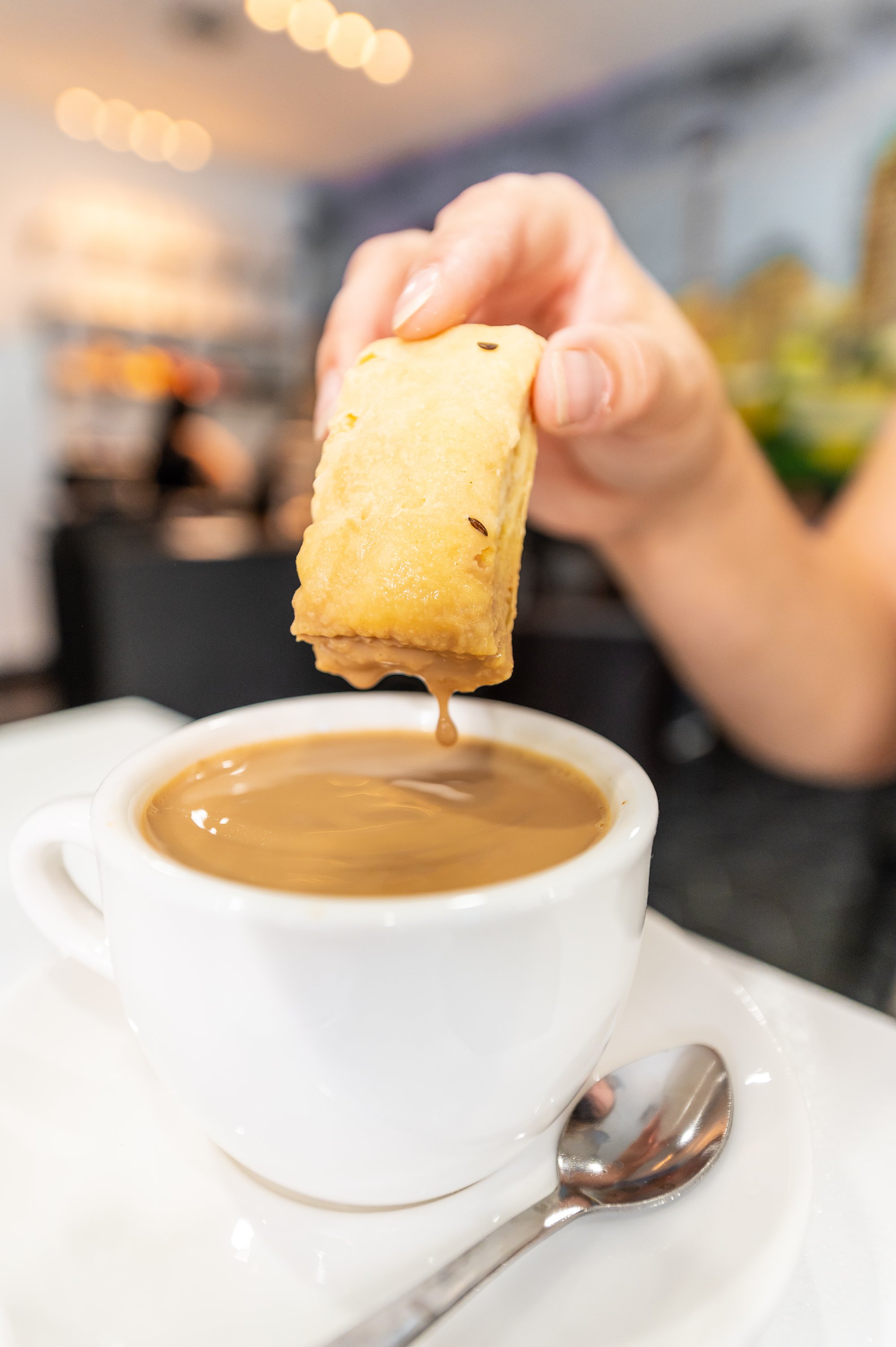 A person's hand holding a churro dipped in hot chocolate at a café. The cup of hot chocolate is on a saucer, with a spoon beside it. The background is blurred with warm lighting.